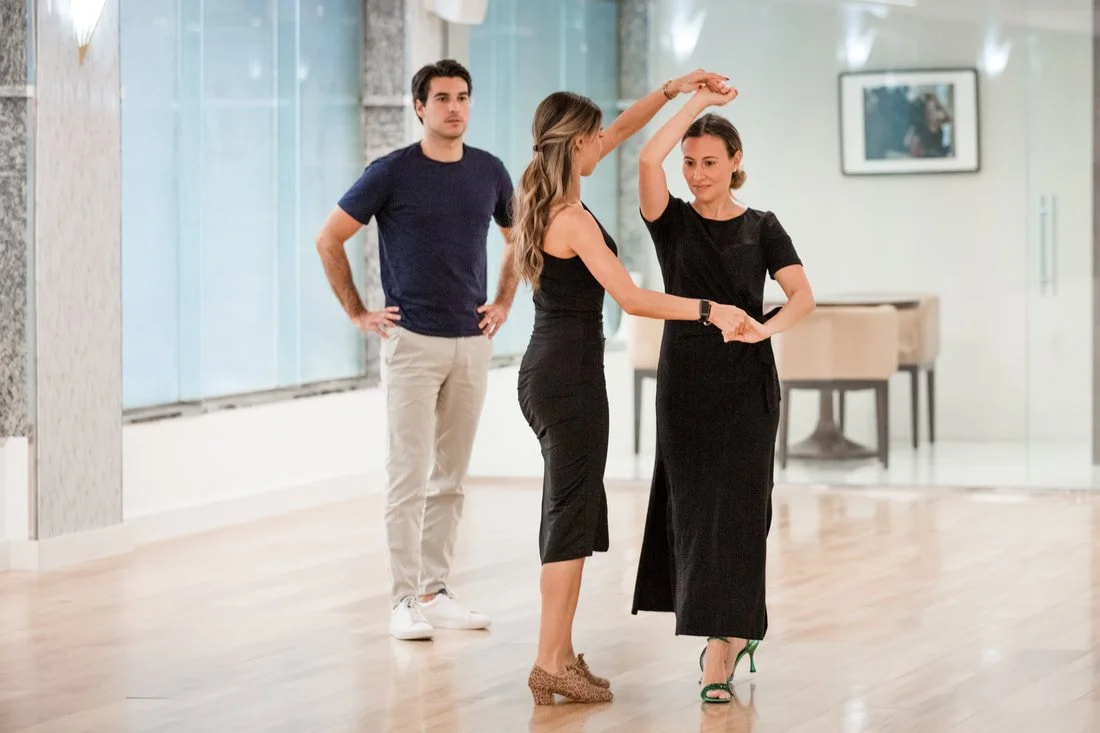 Three people dance in a bright dance studio with wooden floors and large windows. Two women are dancing together, holding hands, one with a raised arm, and the man watches nearby.