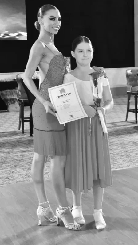 A woman and a young girl standing on a dance floor at an award ceremony. The woman is holding a certificate and both are holding a rose. The woman is dressed in a sparkly dress and heels, while the girl wears a dress and ballet shoes.