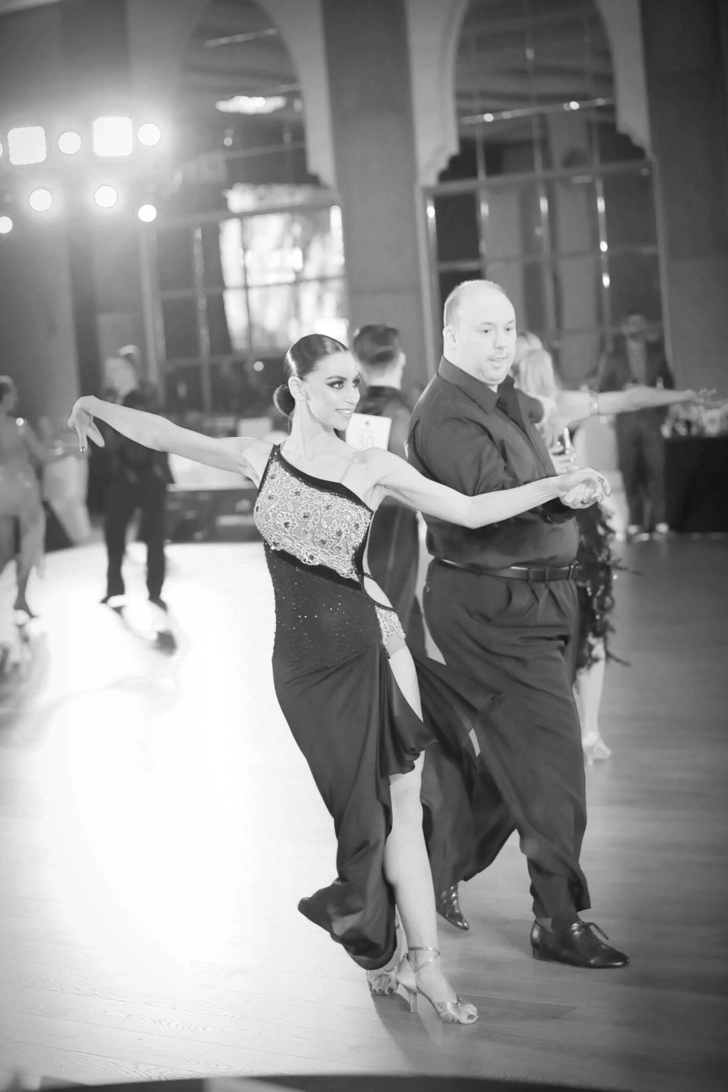 A couple dancing ballroom dance indoors with other dancers in the background, wearing elegant attire, on a polished wooden floor.