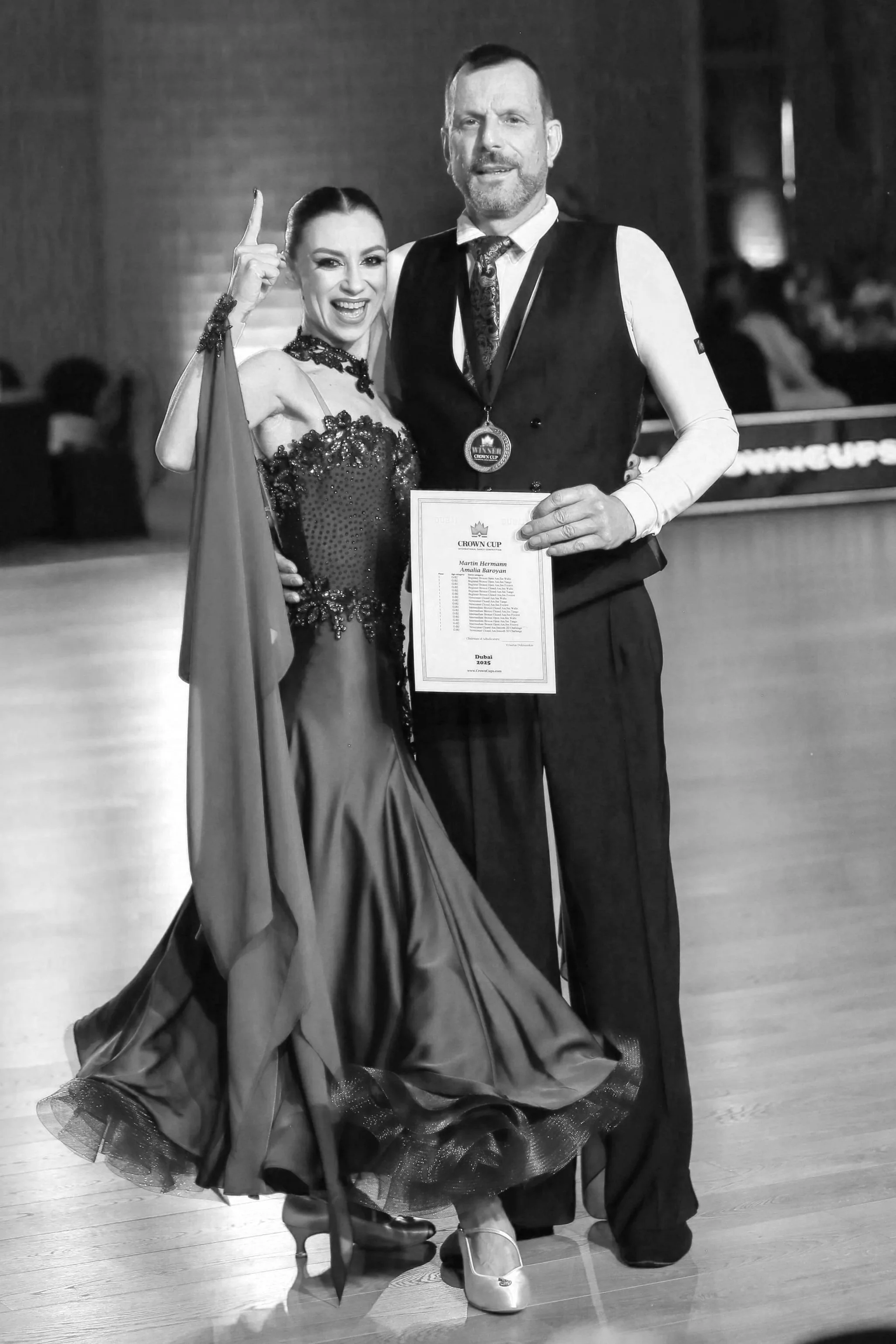 A woman and a man standing together on what appears to be a dance floor. The woman is wearing an elegant evening gown with intricate details, while the man is dressed in a vest, shirt, tie, and is holding a certificate. The woman is smiling and pointing upwards with her index finger. The background is blurred, suggesting an indoor event or ceremony.