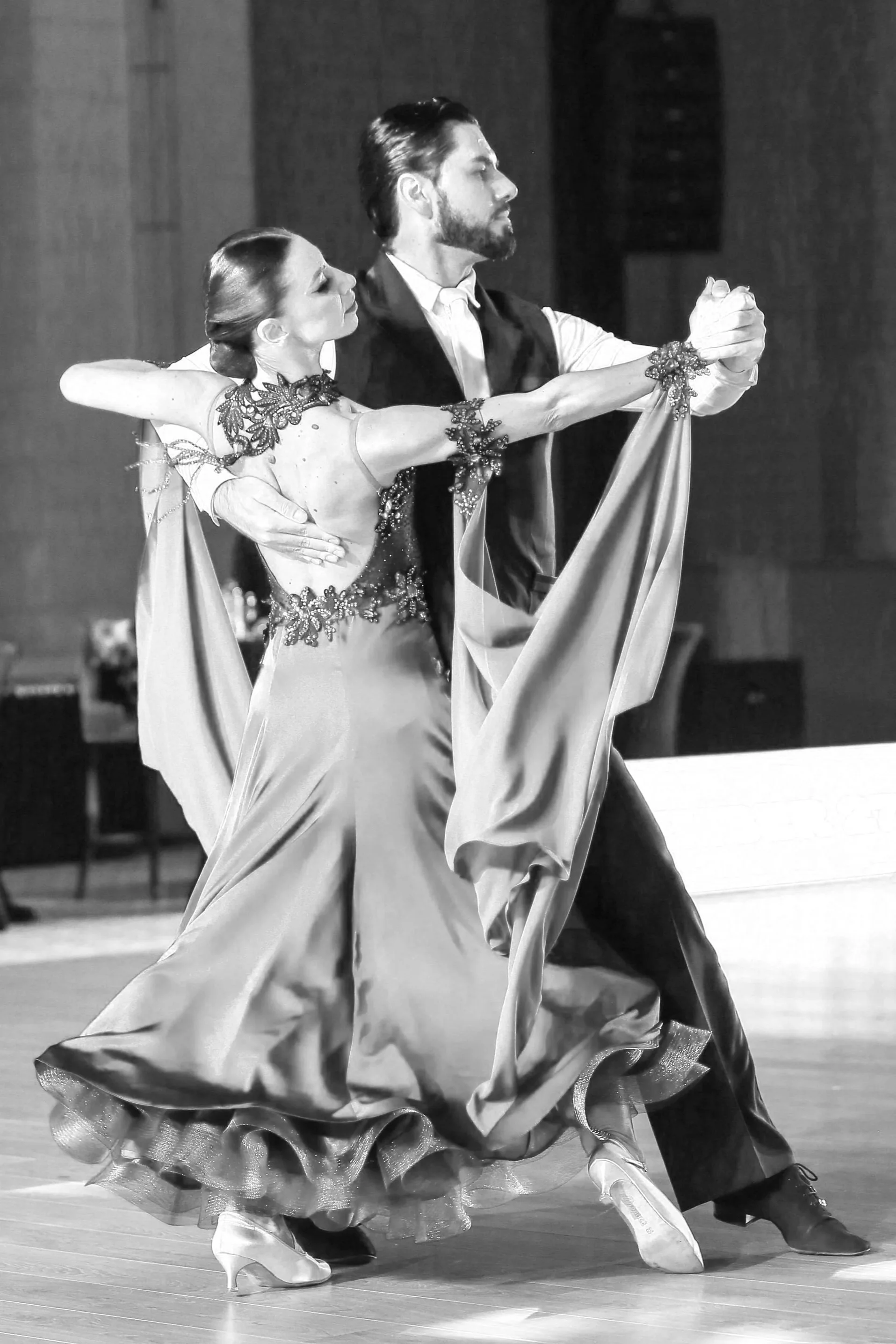 A black and white photo of a ballroom dance couple performing. The woman is in a long flowing dress with detailed embroidery, and the man is in a formal vest and shirt. They are dancing close, with the woman's arm around the man's shoulder and their other hands clasped. The woman is slightly bent forward, and the man is turned to the side with a serious expression.