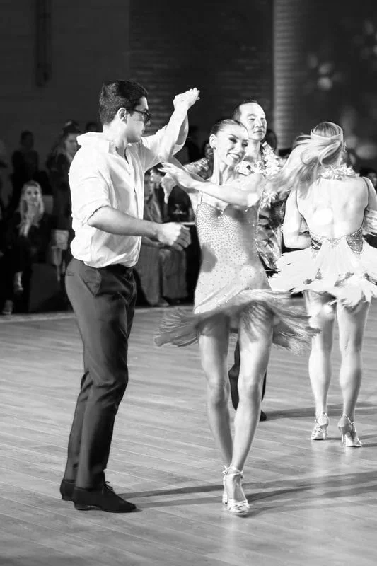 A group of dancers, including a woman in a sparkling dress spinning, participating in a dance competition or performance, with an audience watching in the background.