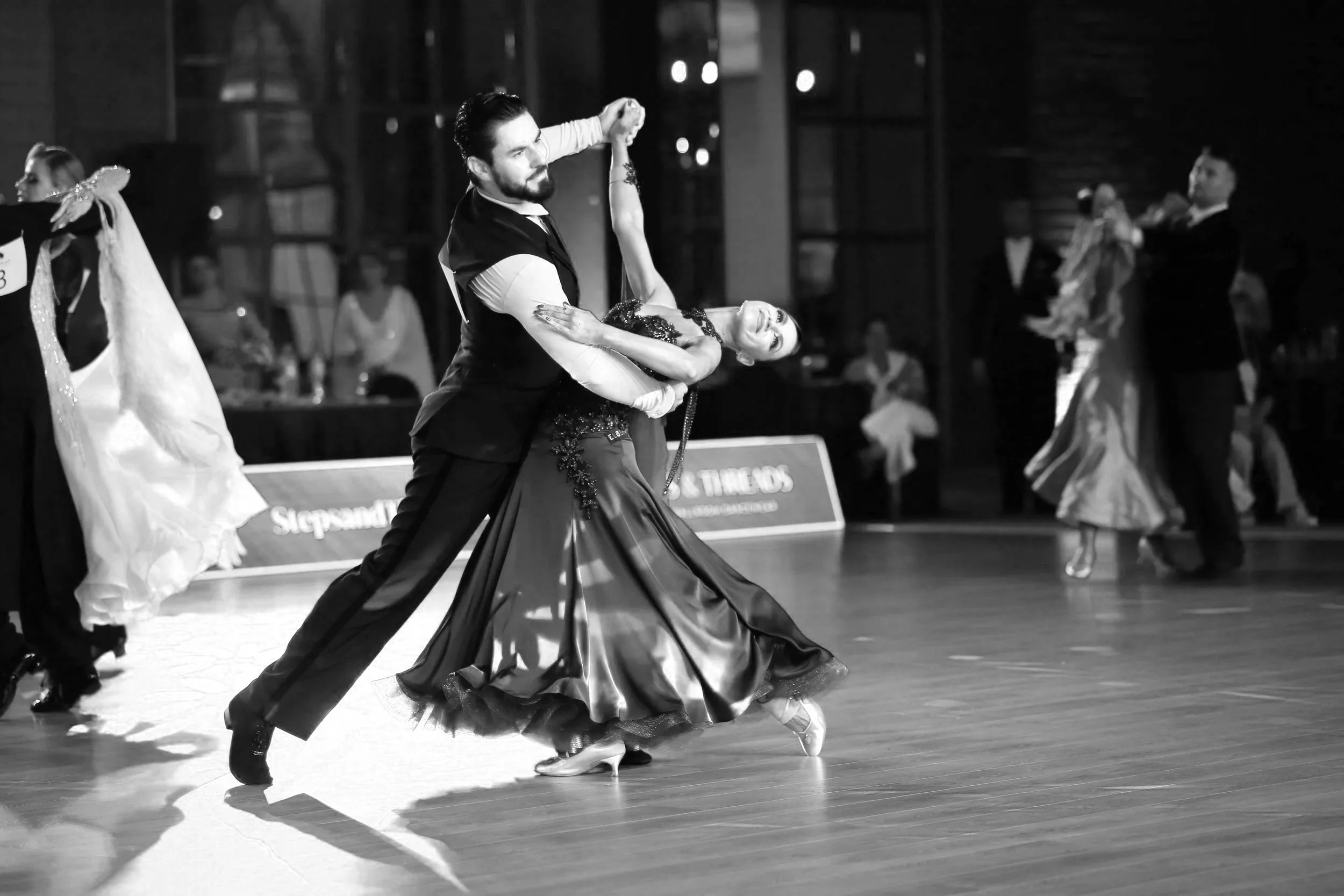 A man and woman dance ballroom in a competition, with other dancers and judges visible in the background, captured in black and white.