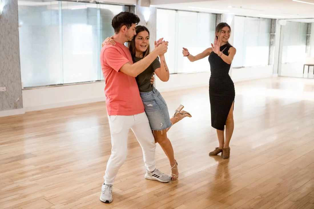 Two people dancing closely with a woman clapping in the background in a large, bright dance studio.