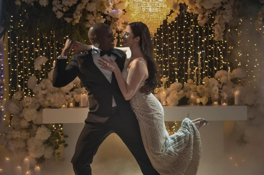 A couple dancing closely at a wedding reception with a decorated backdrop of flowers and fairy lights.