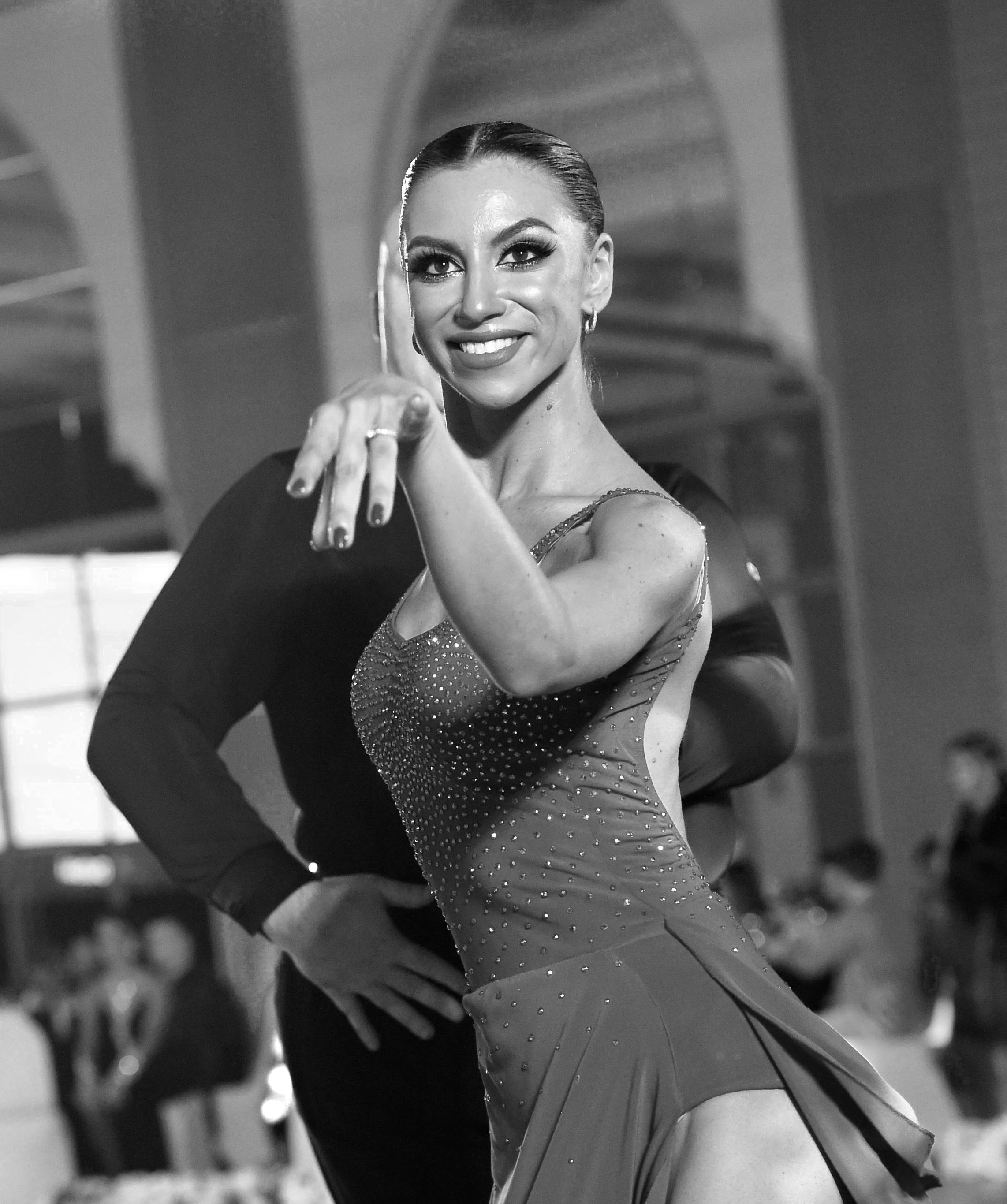 A woman with dark hair, styled in an elegant manner, smiling confidently. She is dressed in a sparkly dance costume, indicating she might be a ballroom or Latin dancer. The background appears to be an indoor venue, possibly a dance hall or stage.