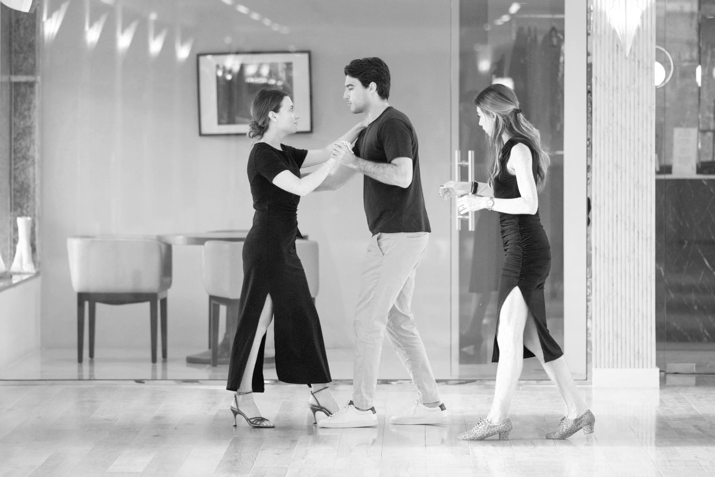 A black-and-white photo of a couple dancing, with a woman in a long skirt and heels, and a man in casual clothing. The woman is holding the man's hand and looking into his eyes, while the man looks at her. A woman with long hair, wearing a sleeveless top and animal-print shoes, is nearby holding a bottle, looking down. The background shows a framed picture on the wall and modern interior decor.