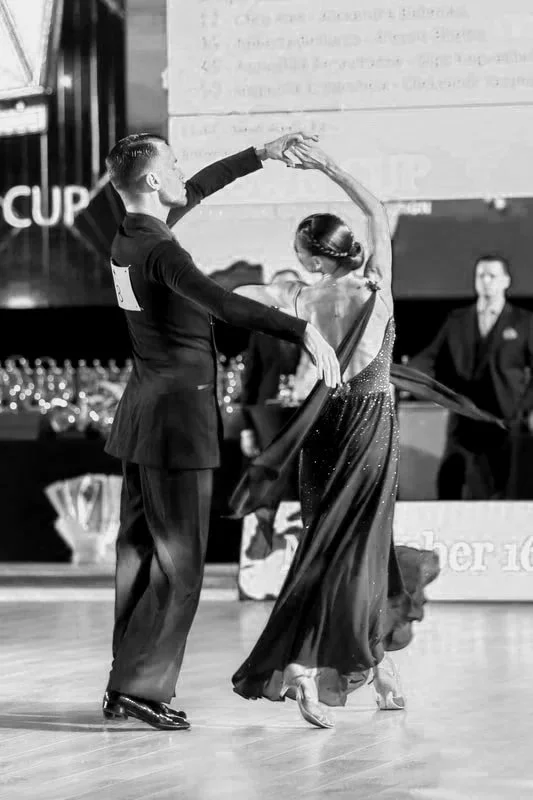 A man and woman dancing ballroom in formal attire at a competition, with an audience and judges in the background.