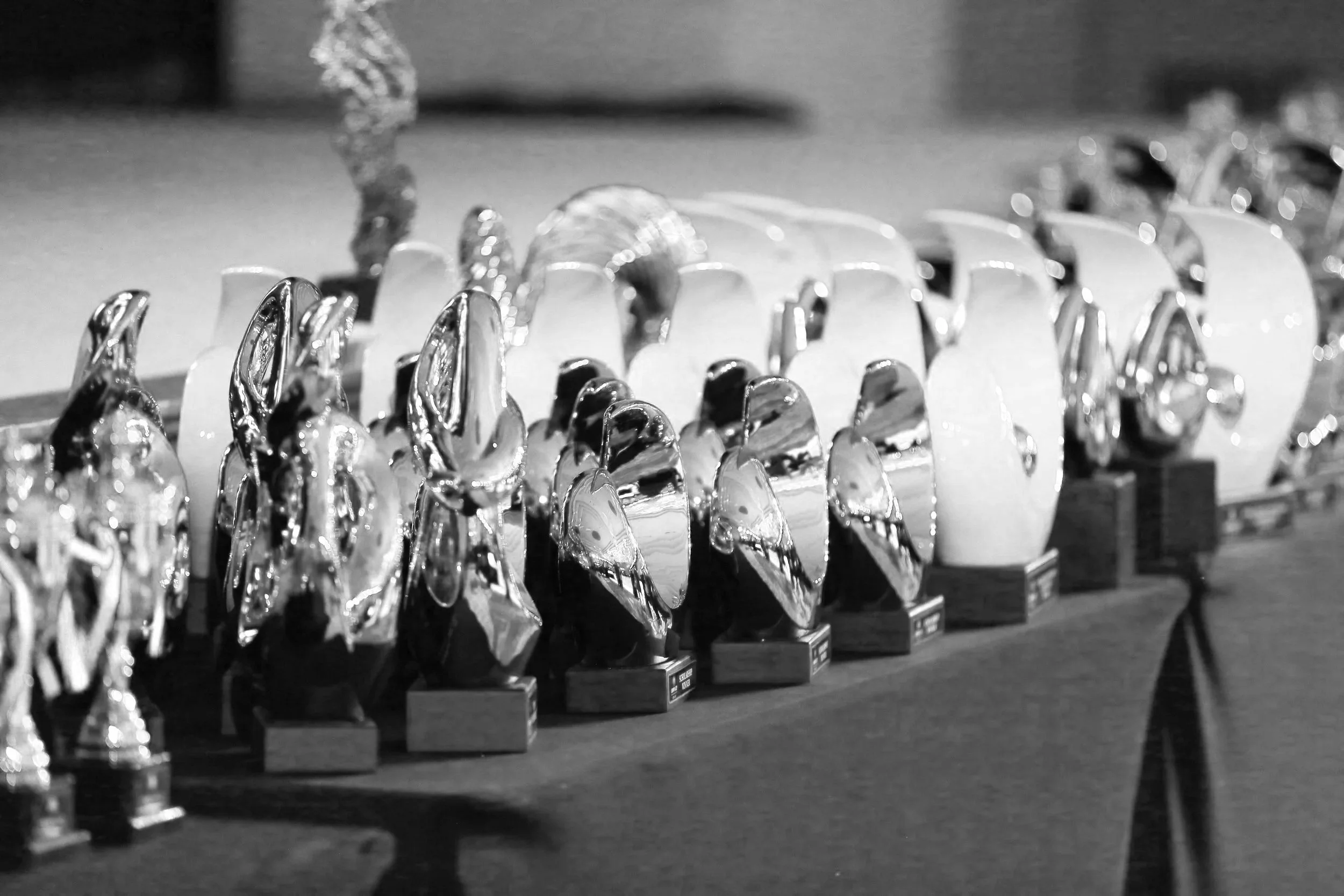 A row of trophies with a sleek, shiny design on display, with each trophy set on a small rectangular base, arranged in a line on a wooden surface, and a blurred background.