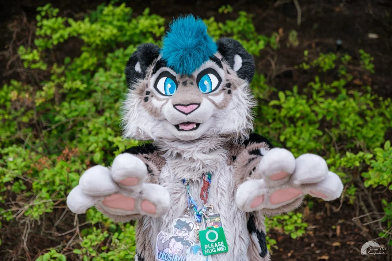 Person in a tiger mascot costume with blue hair, reaching out with both hands, standing outdoors in front of green bushes.