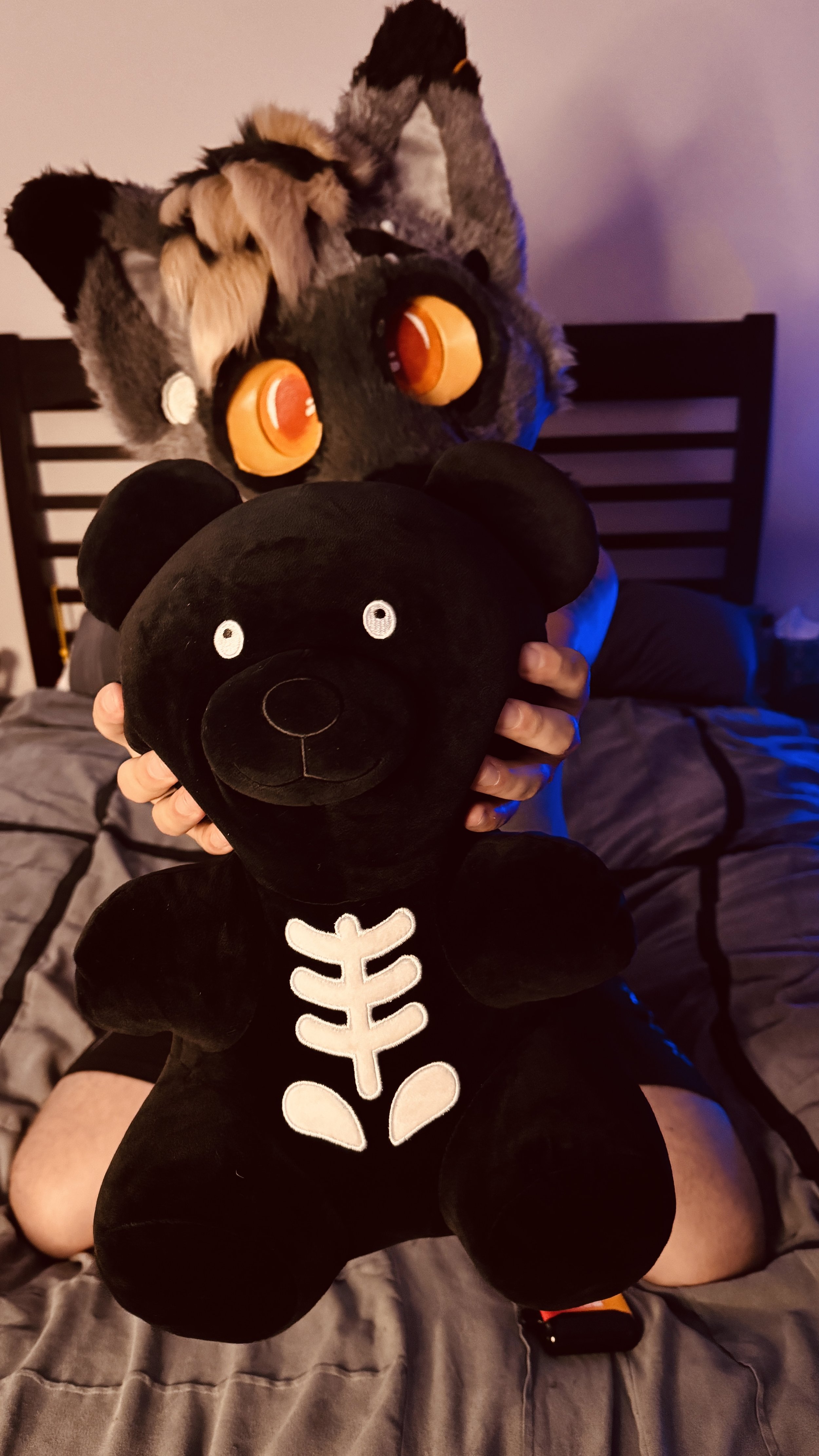 Person laying on bed holding a black plush bear with a white skeleton design on its chest, in front of two large furry animal masks on the bed, one resembling a wolf and the other a raccoon with orange eyes.
