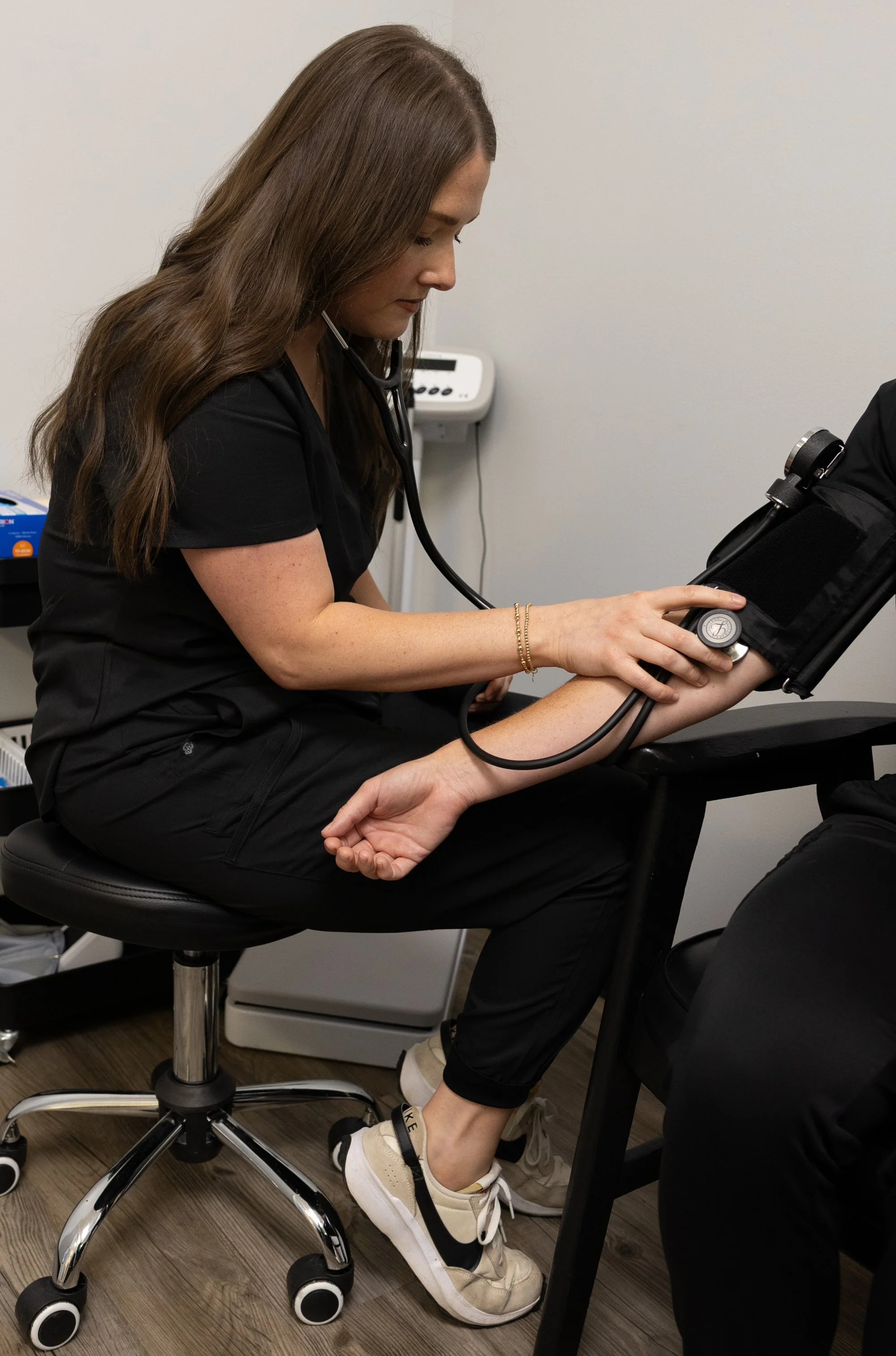 A female healthcare worker in black scrubs and white sneakers seated on an adjustable stool, using a stethoscope to examine a patient in a clinic room.