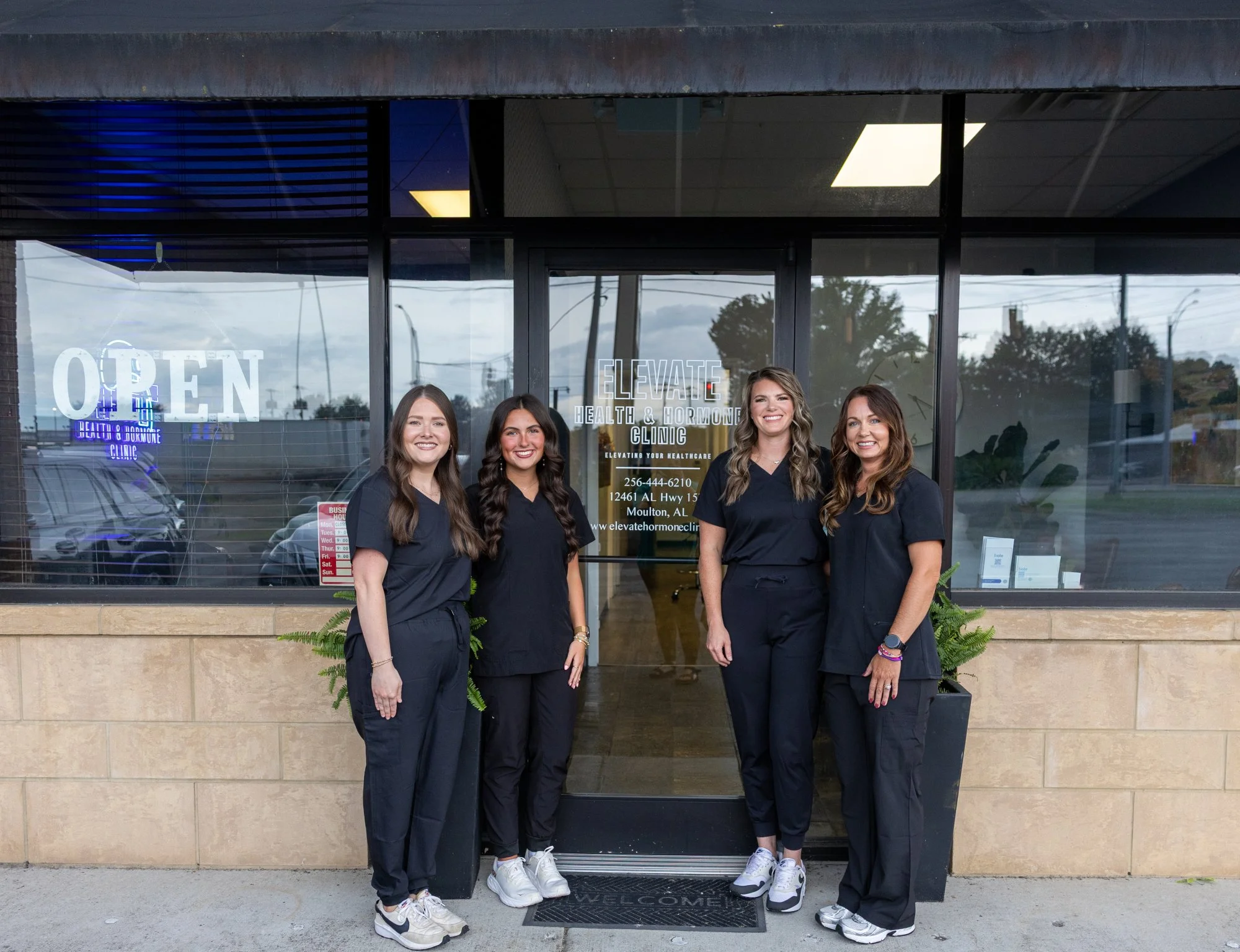 Four women wearing black scrubs standing outside the entrance of a clinic named 'Elevate Health & Hormone Clinic,' smiling for the photo, with glass doors and a sign displaying the clinic name, address, and phone number.