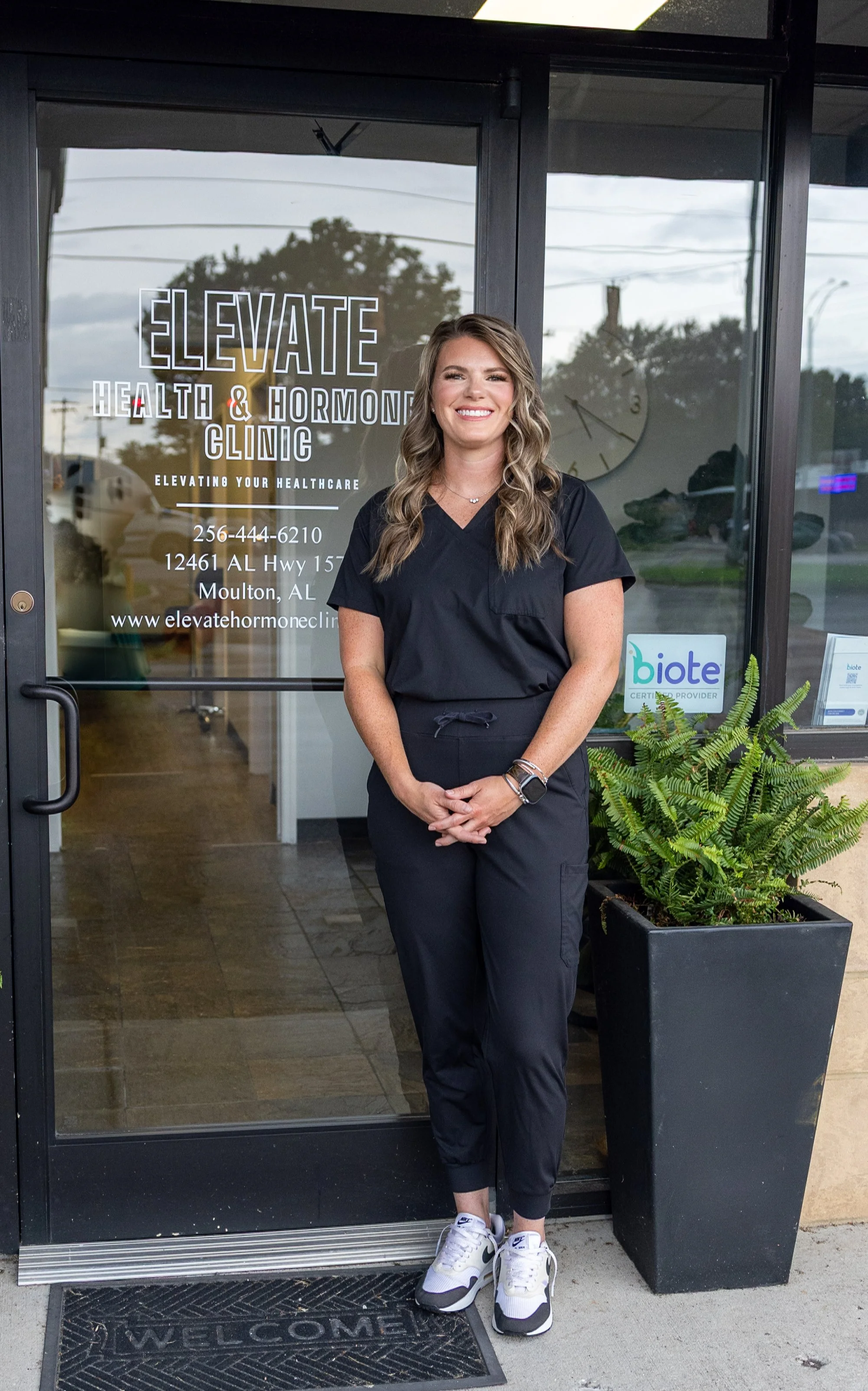 A woman in black scrubs and white sneakers standing outside a healthcare clinic entrance with a glass door. The door has the clinic's name 'Elevate Health & Hormone Clinic' and contact information displayed. A potted green fern is next to her, and a welcome mat is on the ground. The woman is smiling and has wavy brown hair.