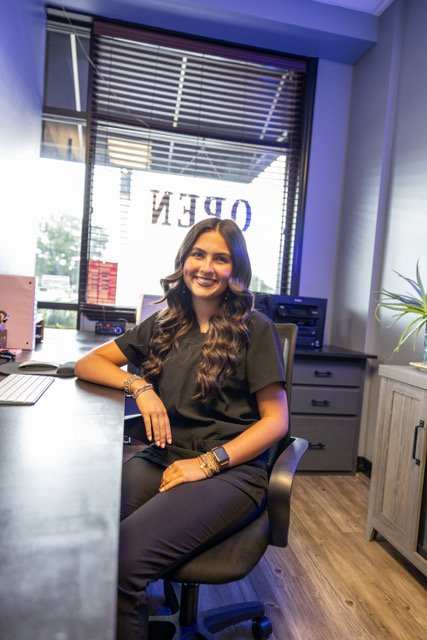 A woman with long, wavy brown hair, wearing a black uniform, sitting at a desk in an office, smiling at the camera.