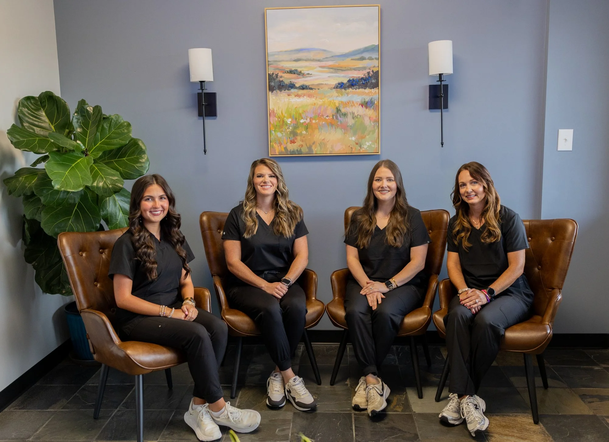 Four women in matching black scrubs sitting on brown leather chairs in a room with a blue wall, two sconces, a colorful landscape painting, and a large green plant.