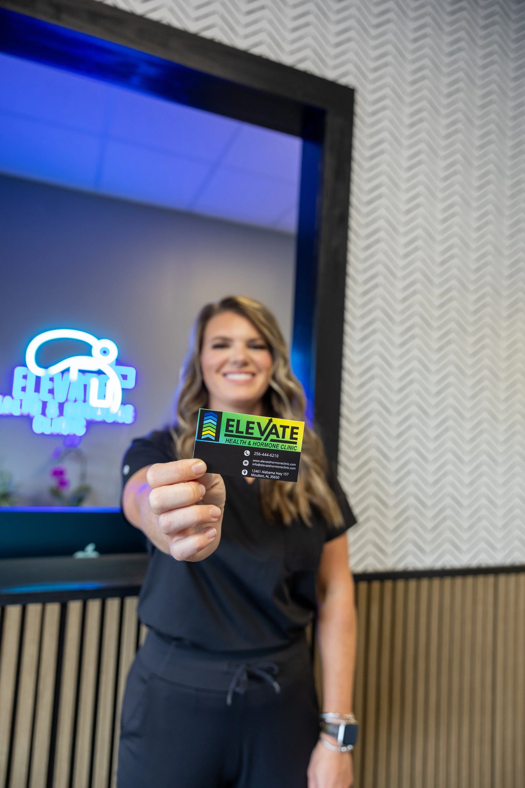 A smiling woman in black scrubs holding a business card for Elevate Health & Hormone Clinic, with a reception area in the background featuring a neon sign and textured wall.