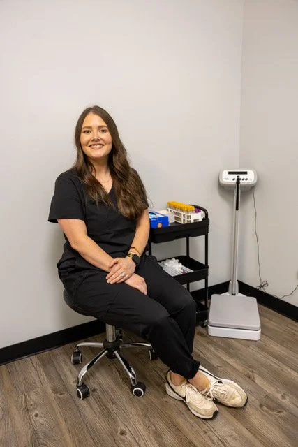 A woman with long brown hair, wearing black scrubs and sneakers, sitting on a rolling stool in a medical or clinical room. She is smiling and sitting next to a small black cart with medical supplies, with a scale in the background.
