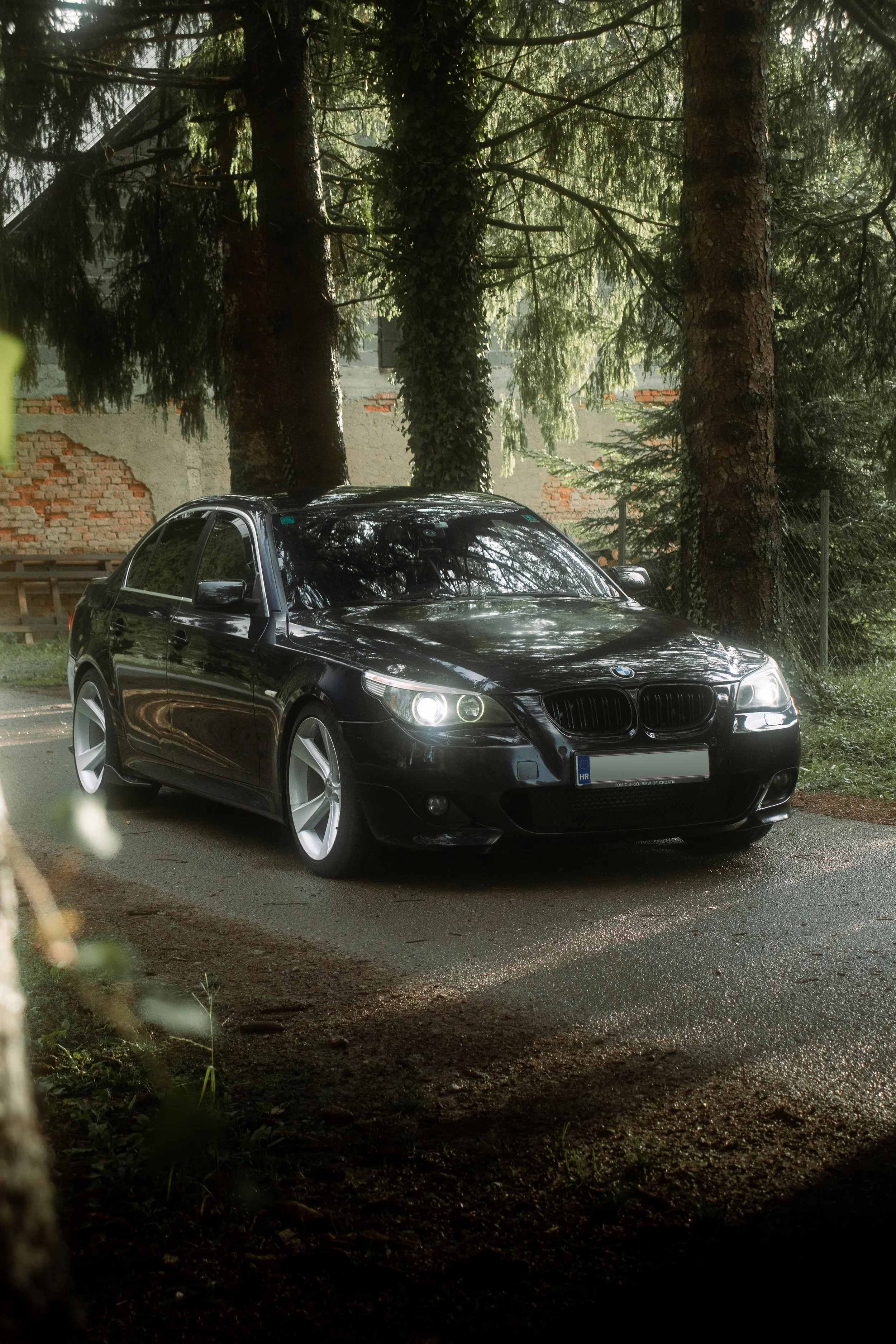 Profesionalna fotografija prednjeg tročetvrtinskog kuta crnog BMW-a E60 s uključenim svjetlima, snimljena na šumskom putu u Hrvatskoj.