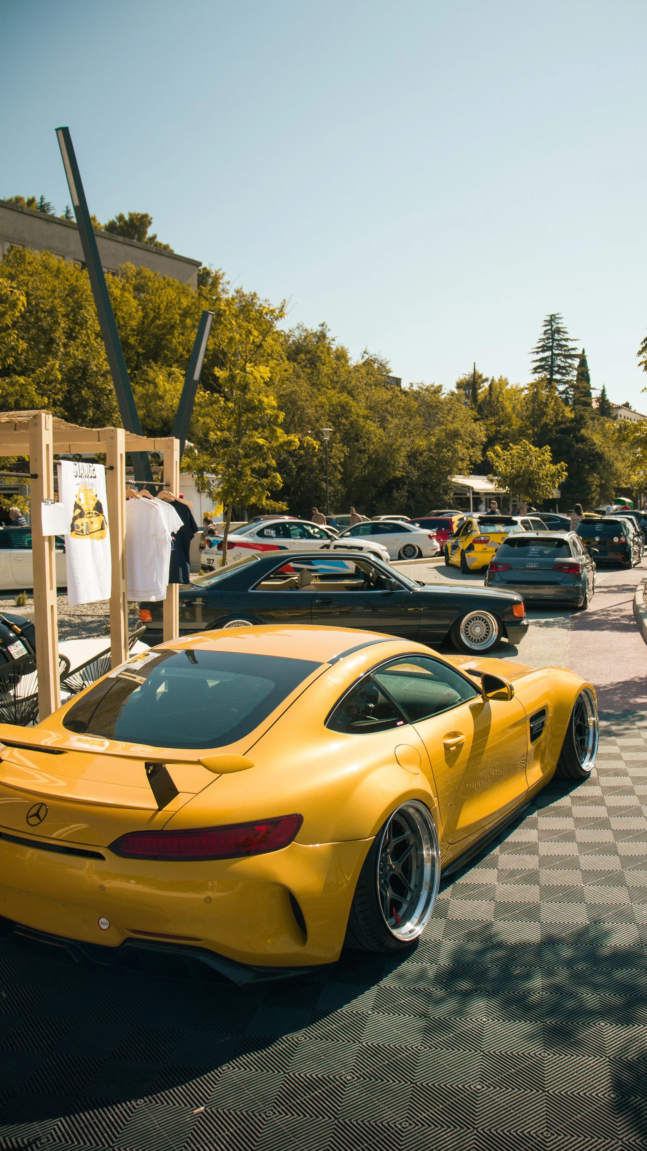 Stražnji bočni kut profesionalne fotografije žutog Mercedes GT-a na kariranoj podlozi tijekom automobilskog eventa, prikazujući styling i kvalitetu izvedbe.