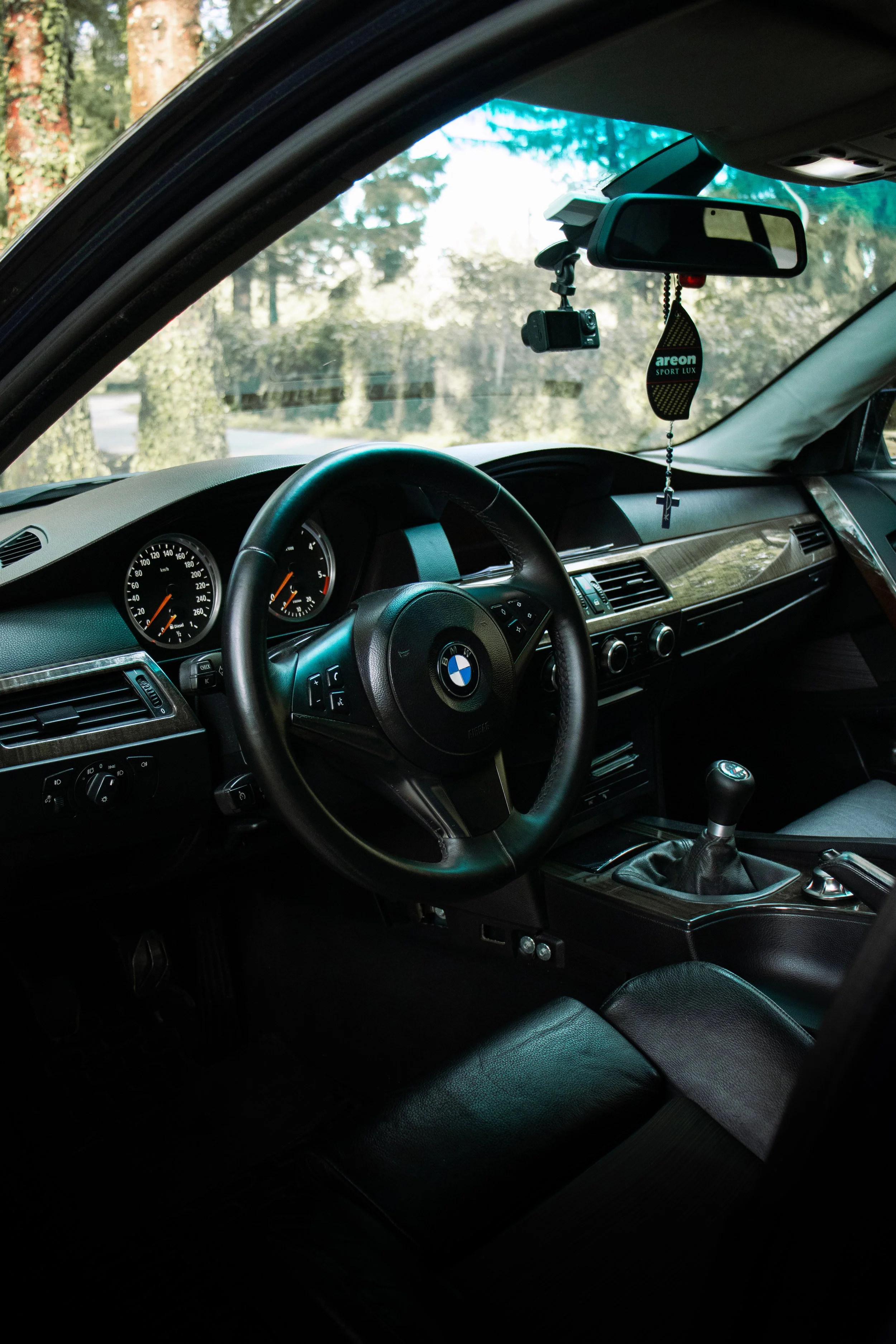 Profesionalna fotografija širokog kuta interijera BMW e60, koja pokazuje pregled kontrolne ploče, upravljača, mjenjača i detalja retrovizora, snimljena s vozačevog sjedala.