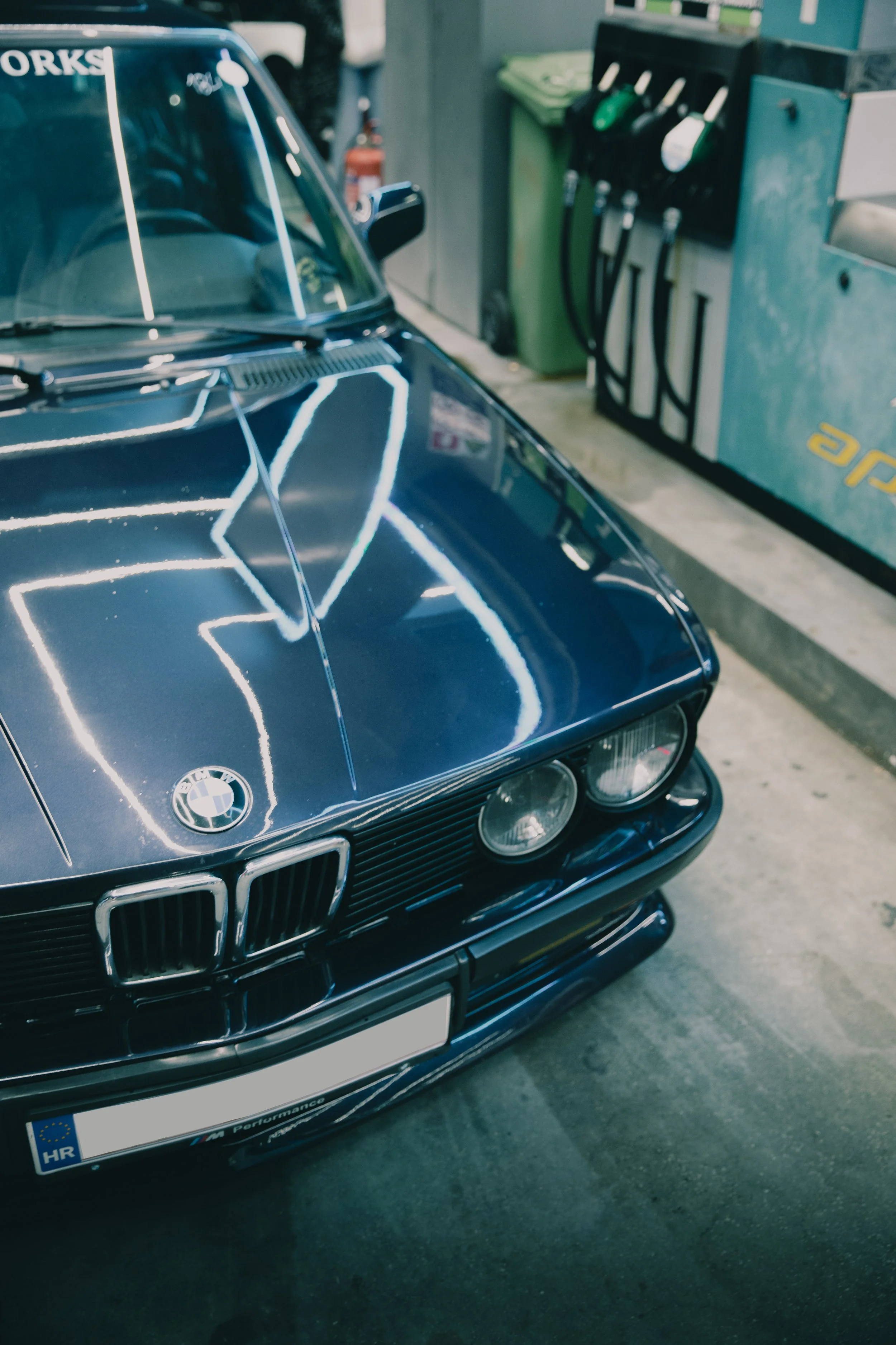 Krupni plan prednjeg kraja i sjajnog poklopca motora tamnoplavog klasičnog BMW-a E28 pod neonskim svjetlima benzinske crpke.