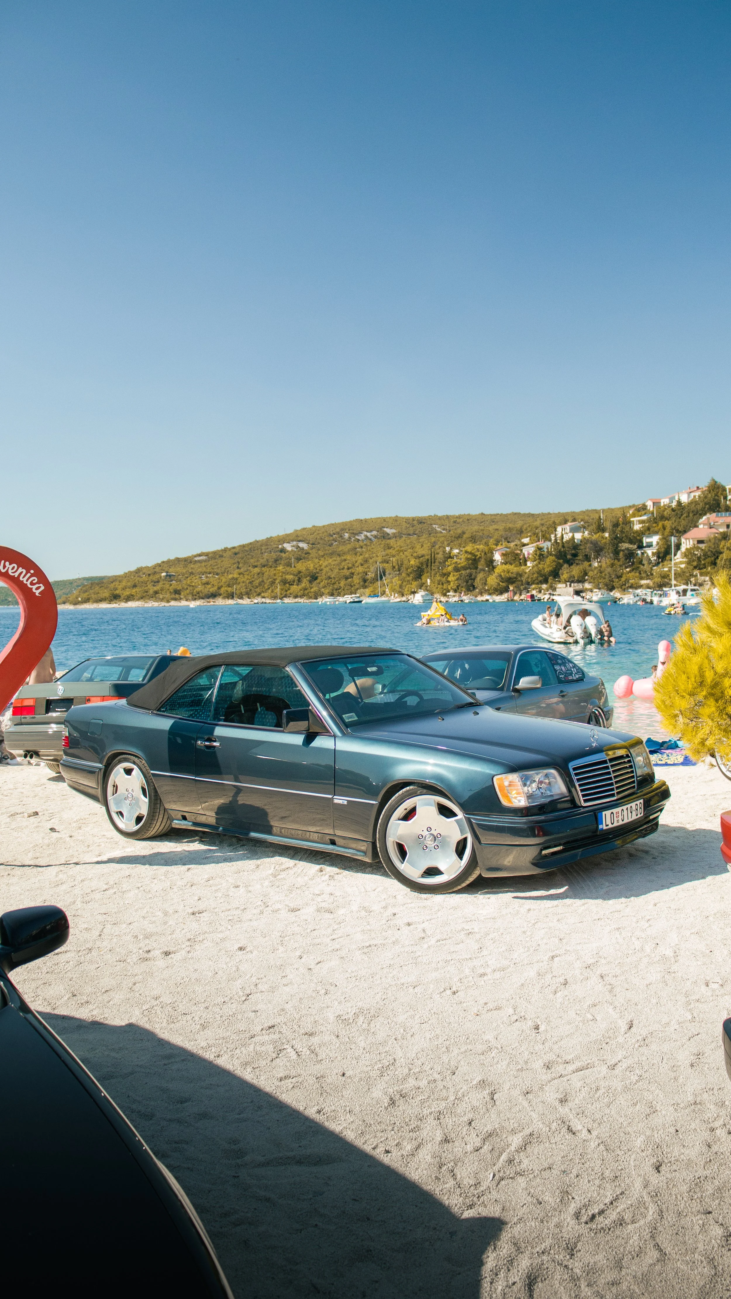 Tamnozeleni klasični Mercedes-Benz W124 kabriolet s Lorinser/AMG naplatcima parkiran na plaži pokraj mora.