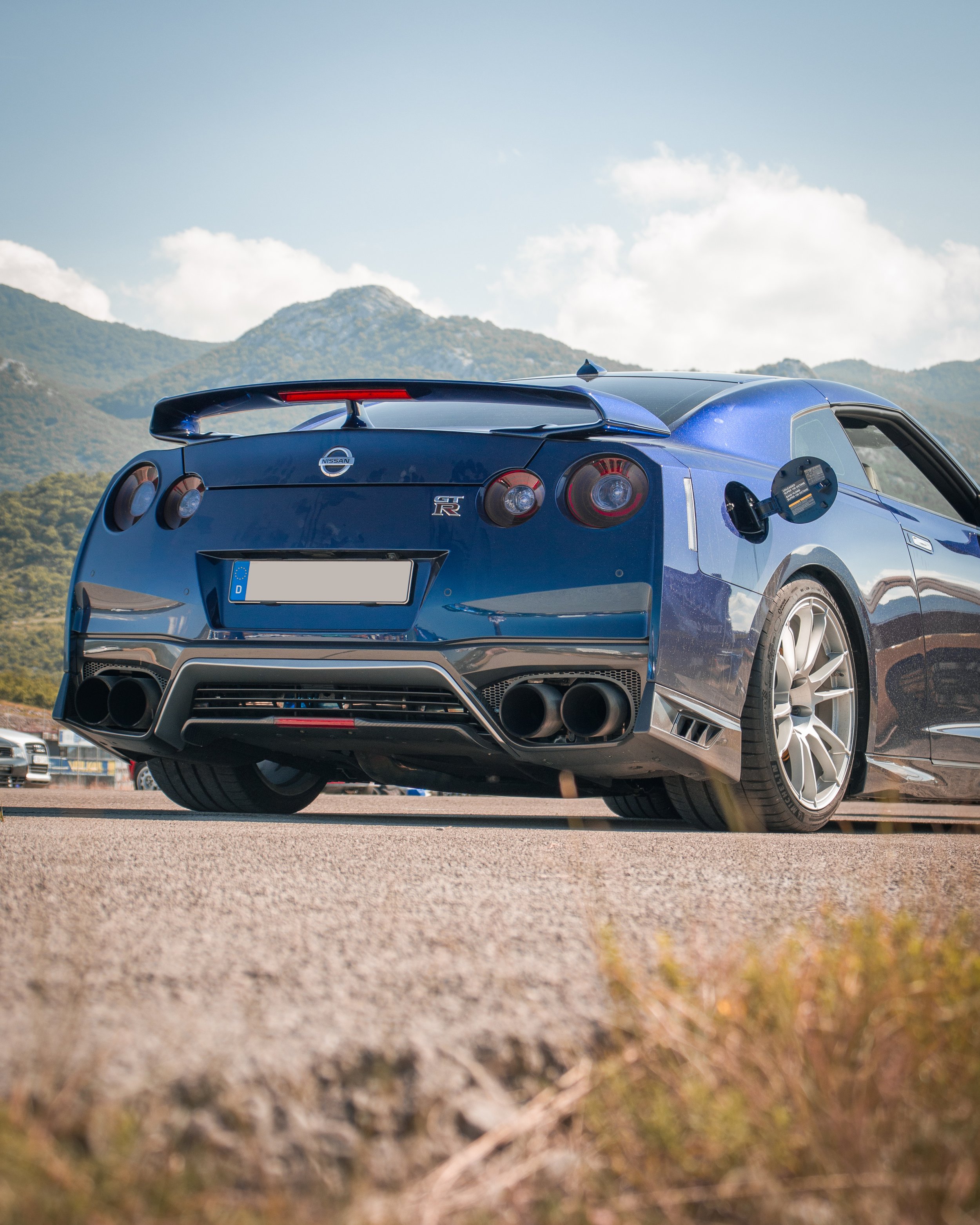 Profesionalna fotografija niskog kuta stražnjeg dijela plavog Nissan GT-R R35 automobila na stazi s planinskom pozadinom u Hrvatskoj.