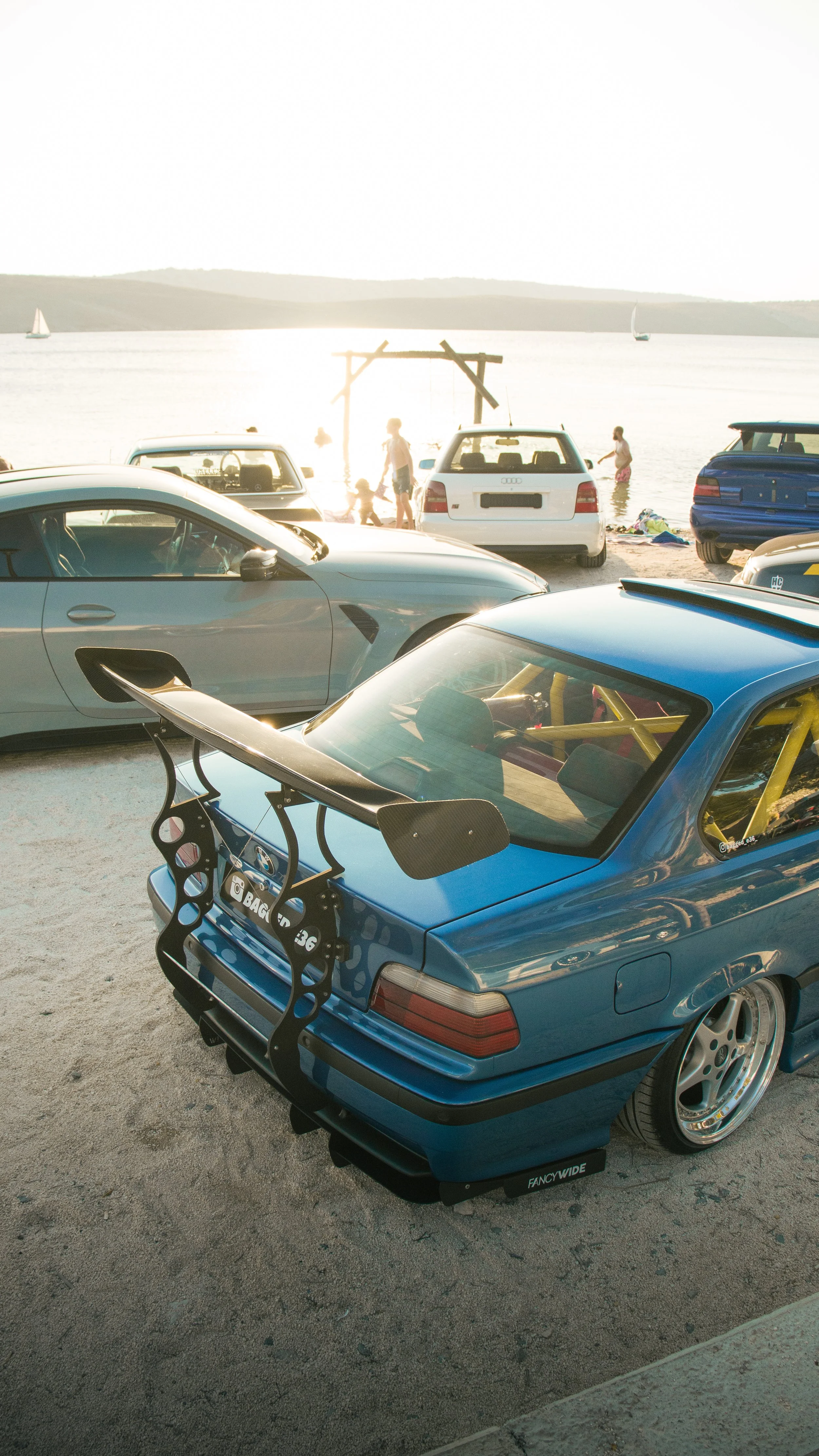 Stražnji kadar plavog BMW-a E36 Coupe s masivnim trkaćim bojler spojlerom i roll barom uz zalazak sunca na plaži.