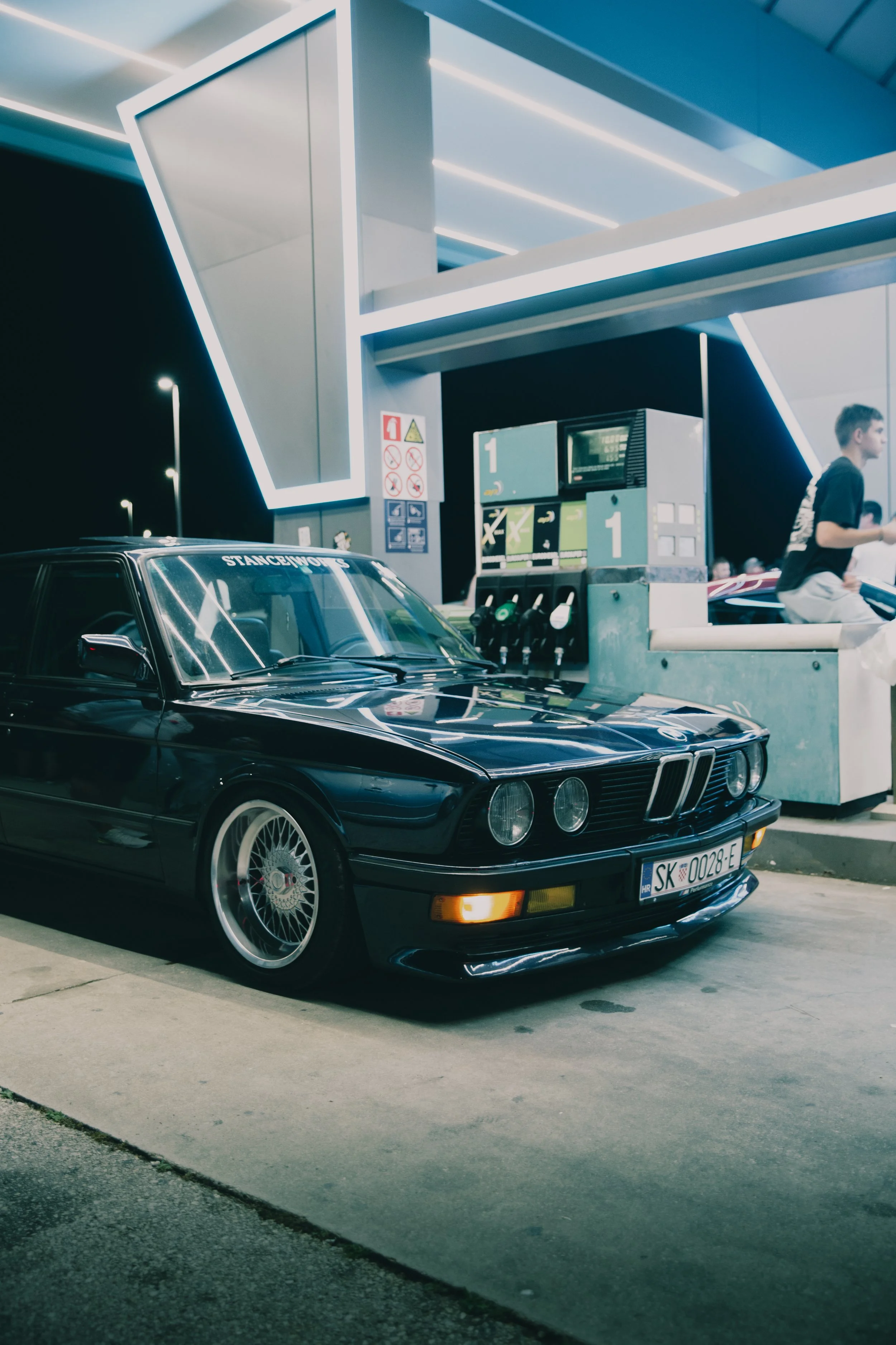 Prednji profil spuštenog klasičnog BMW-a E28 s BBS mrežastim naplatcima i StanceWorks naljepnicom na noćnoj benzinskoj crpki.