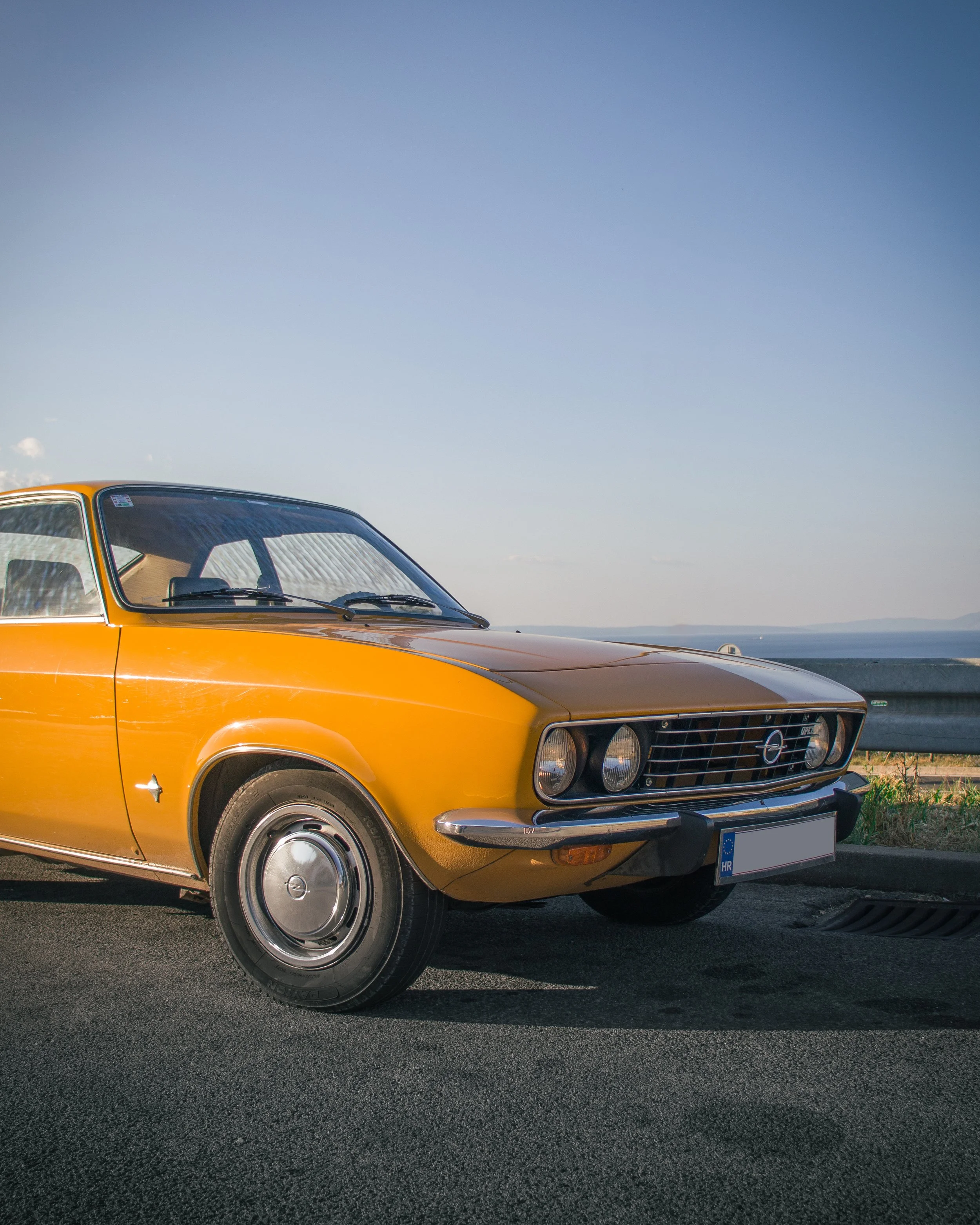 Klasični narančasti Opel Manta Kadett parkiran na otvorenom, pogled sprijeda pod vedrim nebom i morem u pozadini.