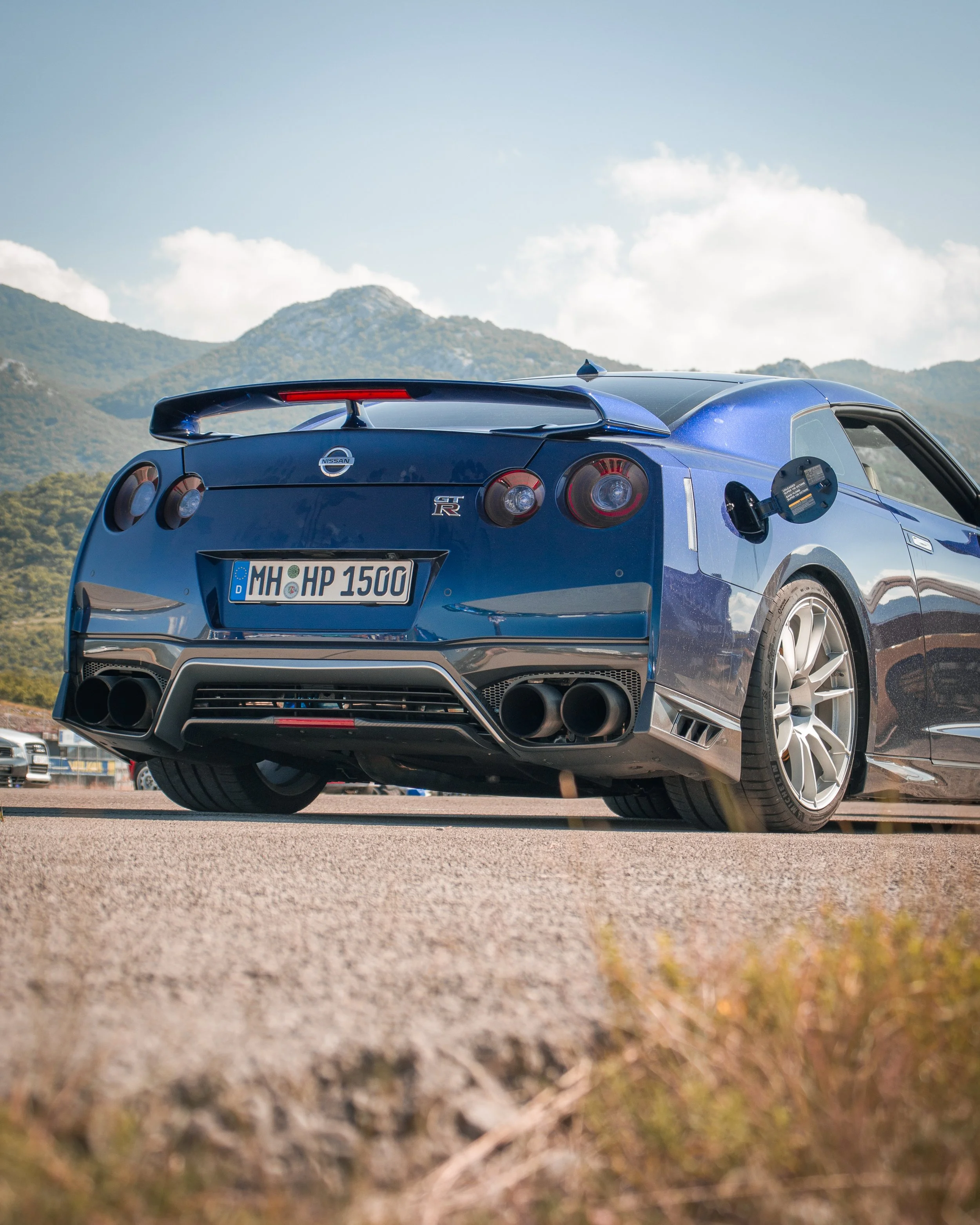Profesionalna fotografija niskog kuta stražnjeg dijela plavog Nissan GT-R R35 automobila na stazi s planinskom pozadinom u Hrvatskoj.