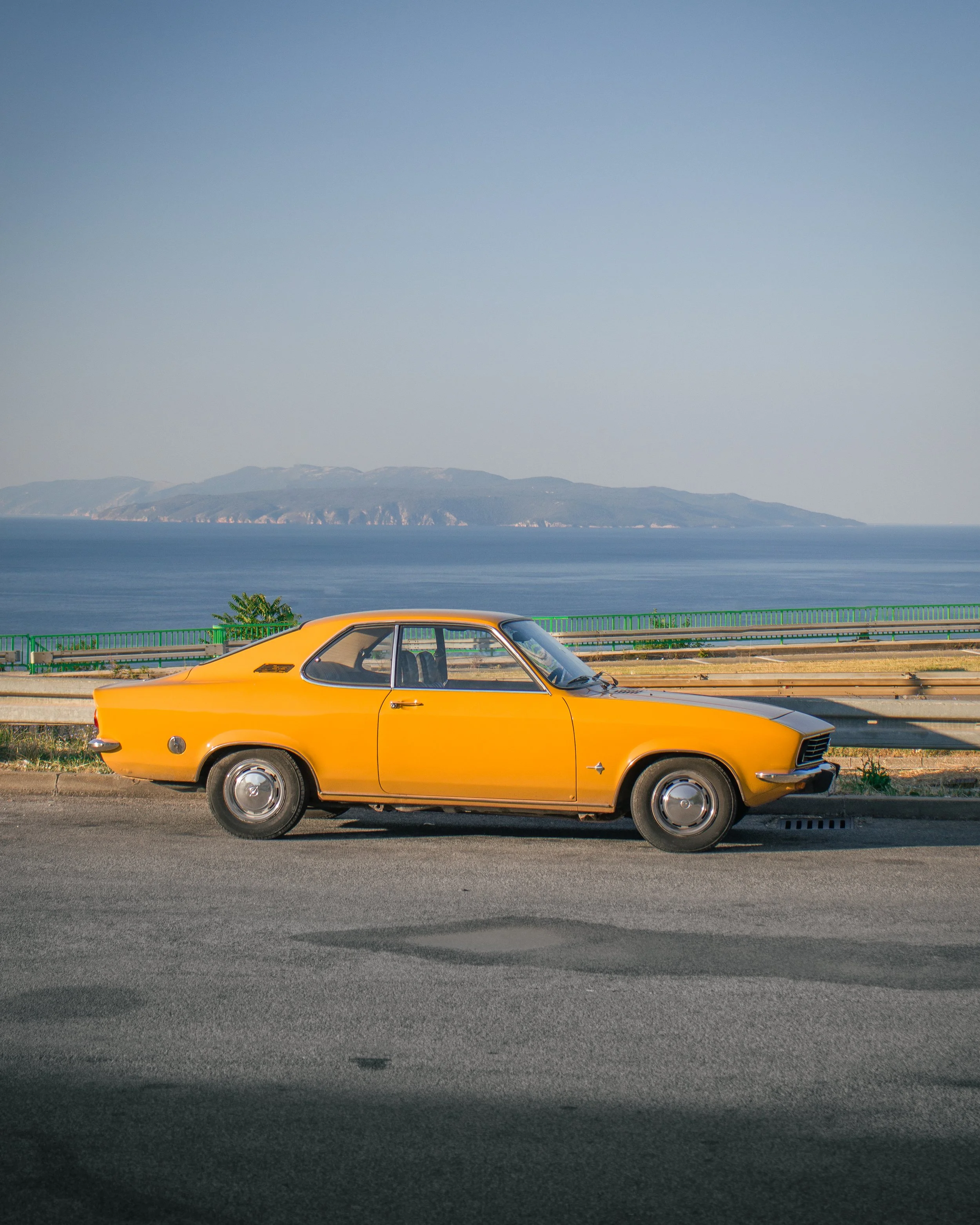 Klasični narančasti Opel Manta Kadett parkiran uz obalu, pogled sa strane s morem i brdima u pozadini