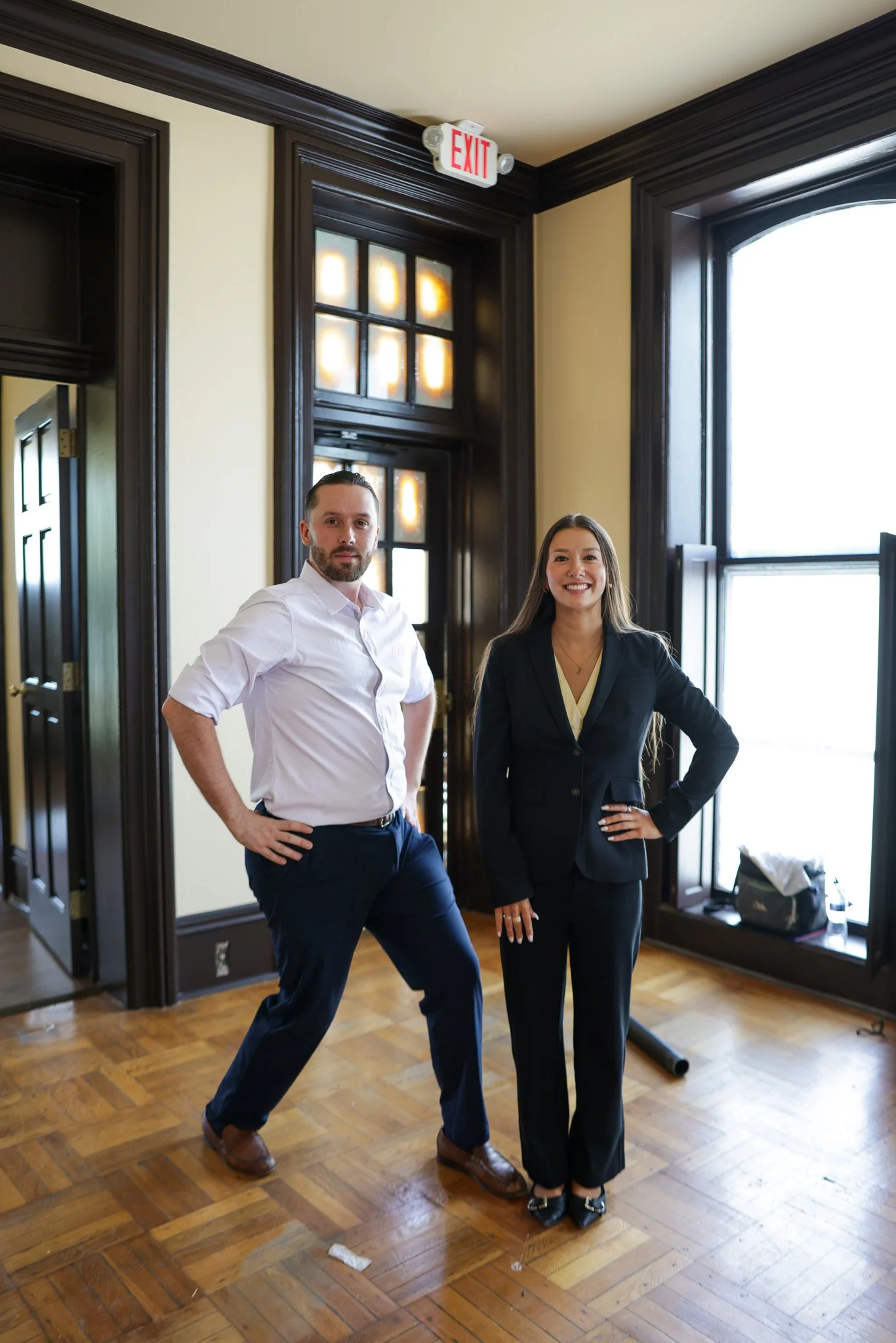 Two professionally dressed individuals, a man and a woman, stand in a room with wooden flooring and large windows. The man is wearing a white button-down shirt and navy pants, while the woman is dressed in a black blazer and matching pants, smiling a