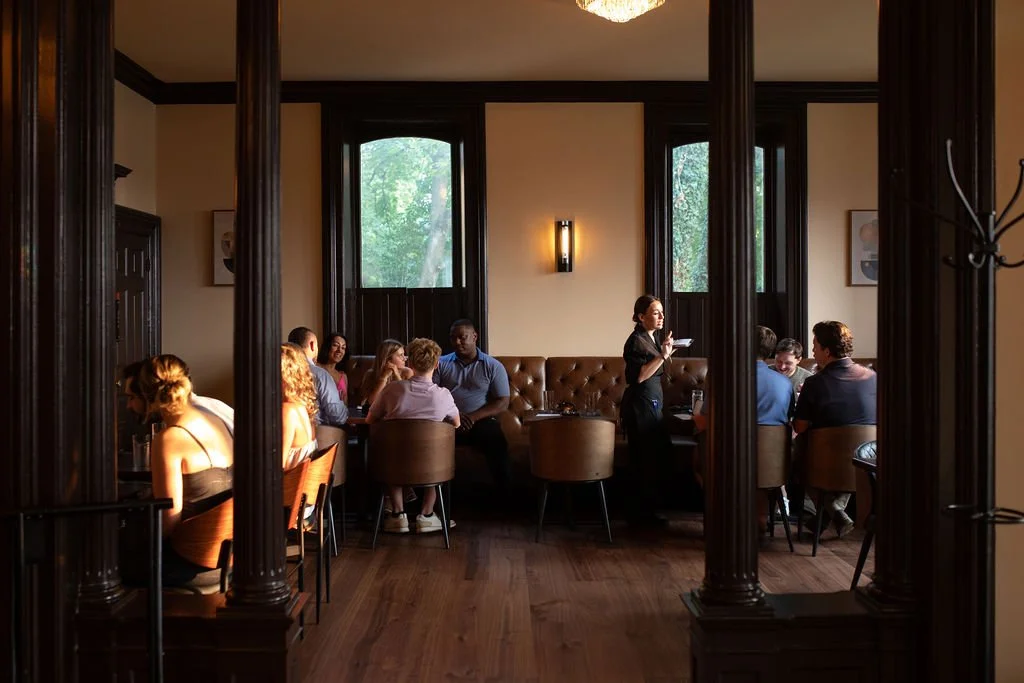 A group of people dining at Table 1888 with large windows, a waitress taking orders, and elegant wooden decor.