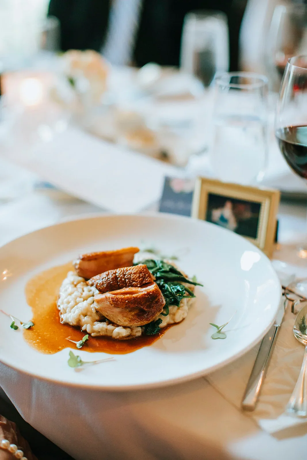 A plated dish with slices of roasted chicken served over creamy rice with sautéed greens and a brown sauce, garnished with microgreens, on a white dinner plate. In the background, there are wine glasses, a small framed photo, and a blurred table setting.