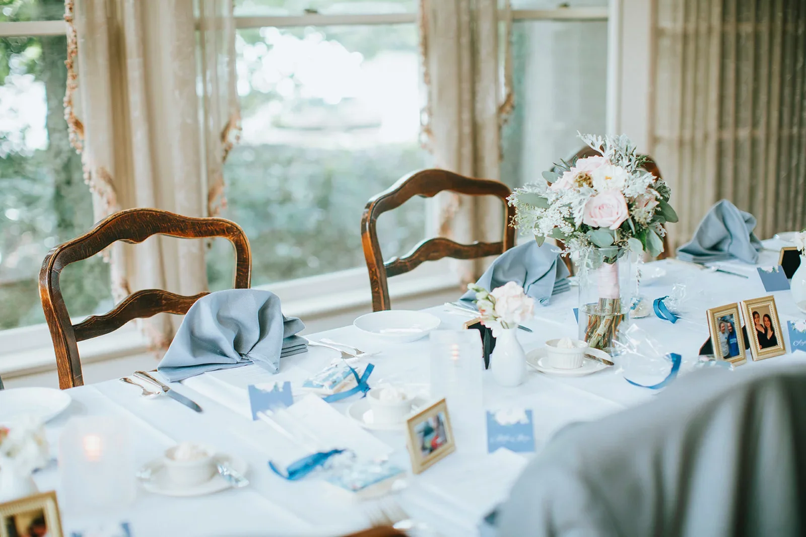 A decorated banquet table set for a wedding reception or celebration, featuring a large floral centerpiece, framed photographs, blue napkins, and elegant tableware, with wooden chairs and a window with curtains in the background.
