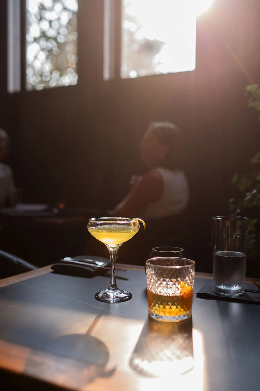 A cocktail glass with a yellow drink on a table, accompanied by a textured glass with an amber beverage and a tall glass of water, in a dimly lit setting with sunlight streaming through windows, and blurred people in the background.