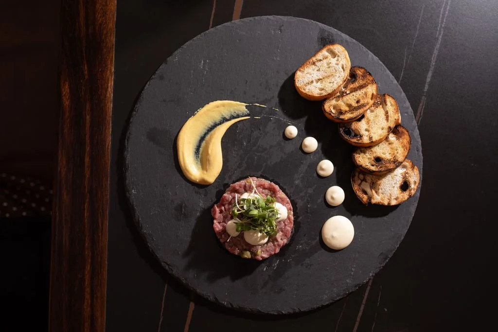 Table 1888 menu item - A black slate plate with grilled bread slices, a serving of tartare topped with microgreens and sauce, and several dollops of creamy sauce.