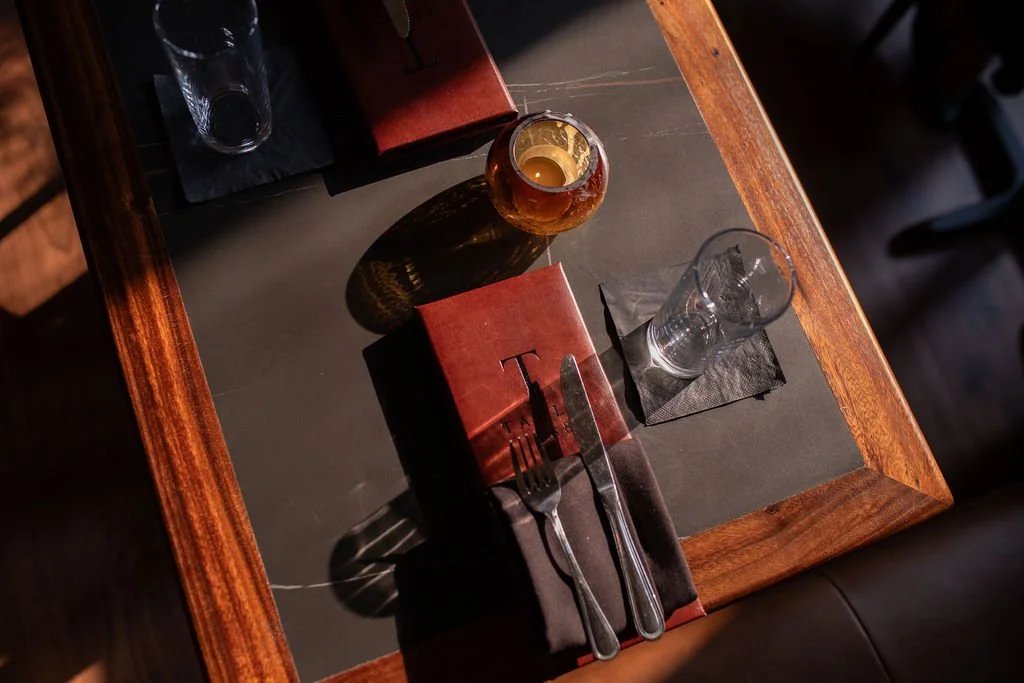 A table setting with a water glass, a candle holder, a red napkin with a fork and knife on top, and a small red menu card, with sunlight casting shadows.