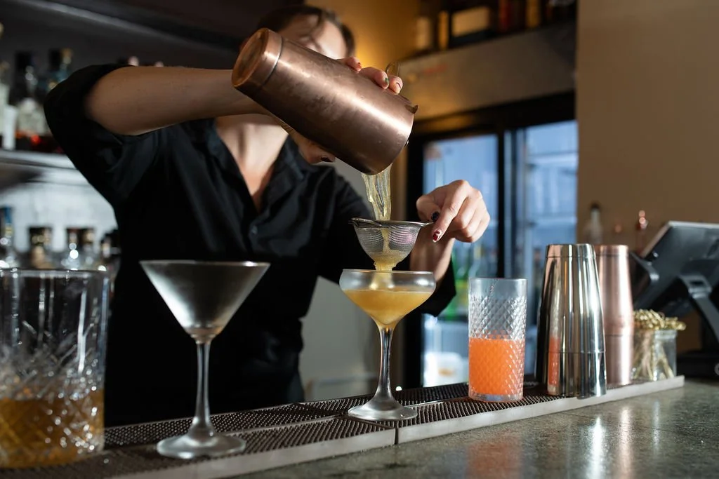 A bartender pouring a yellow cocktail through a strainer into a coupe glass at a bar.