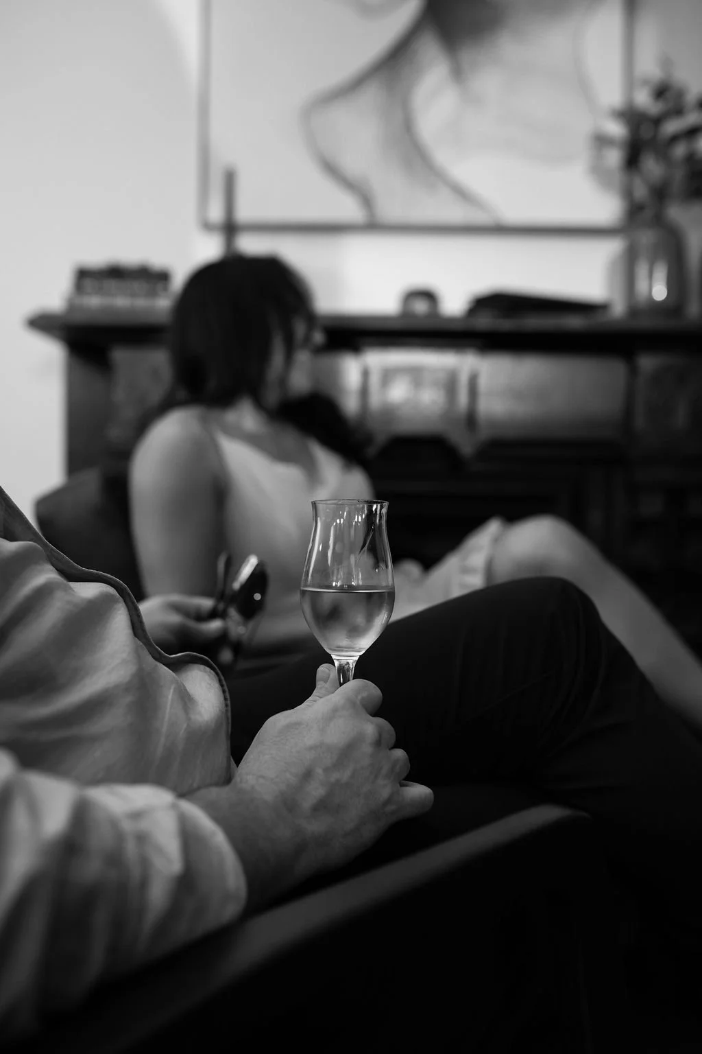 A person holding a glass of water or clear beverage in a living room, with a woman in the background sitting and using her phone, in a black and white photo.