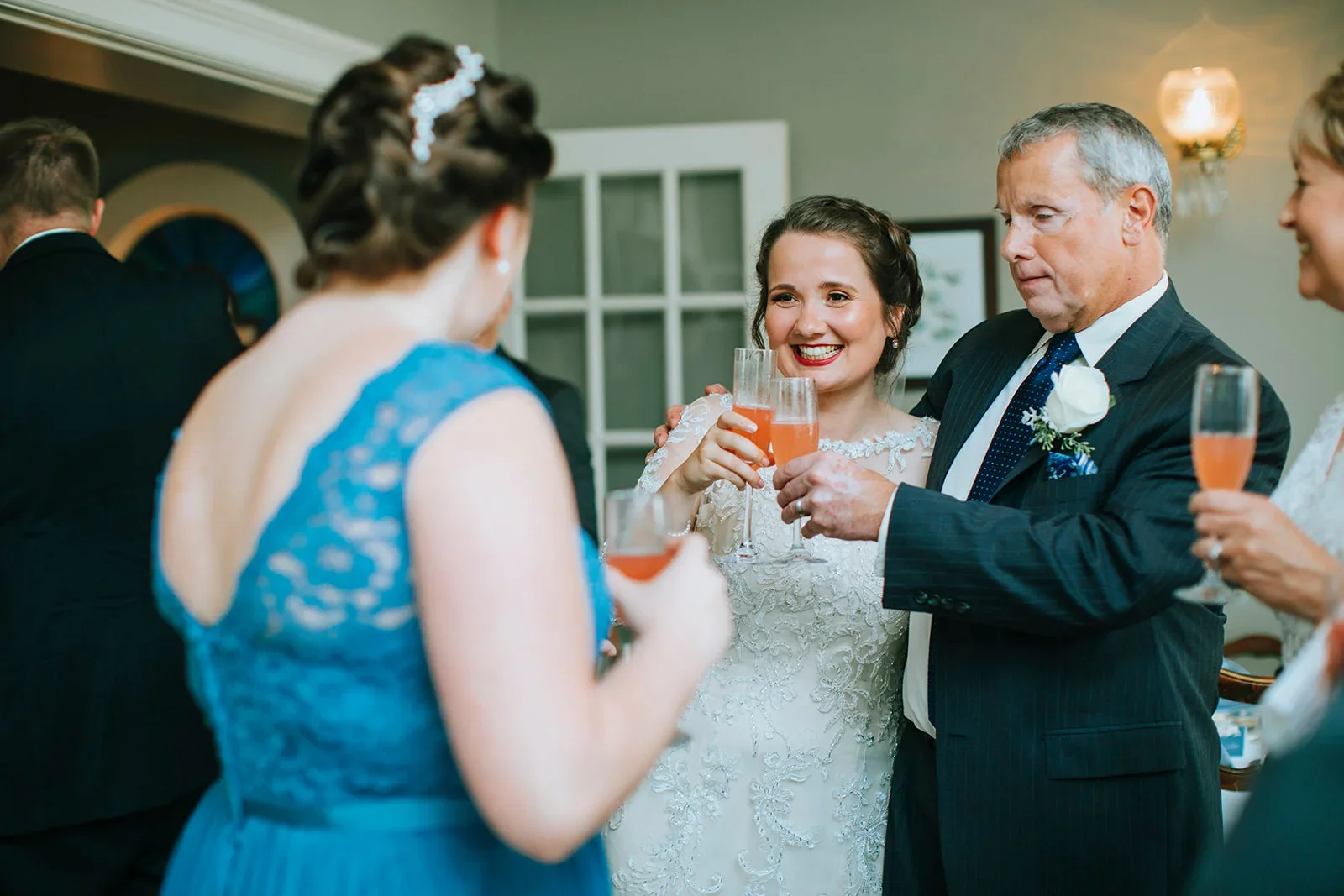 Group of people celebrating at a wedding reception, raising champagne glasses, including a bride in a white lace dress and an older man in a suit with a white boutonniere.