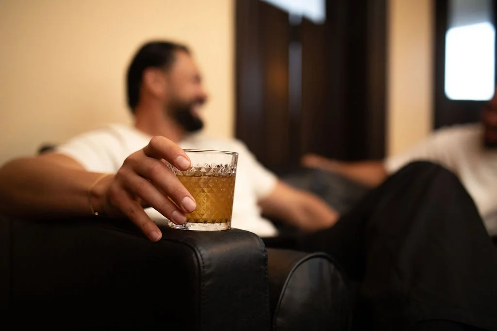 Person holding a glass of whiskey while sitting on a couch with another person in the background.