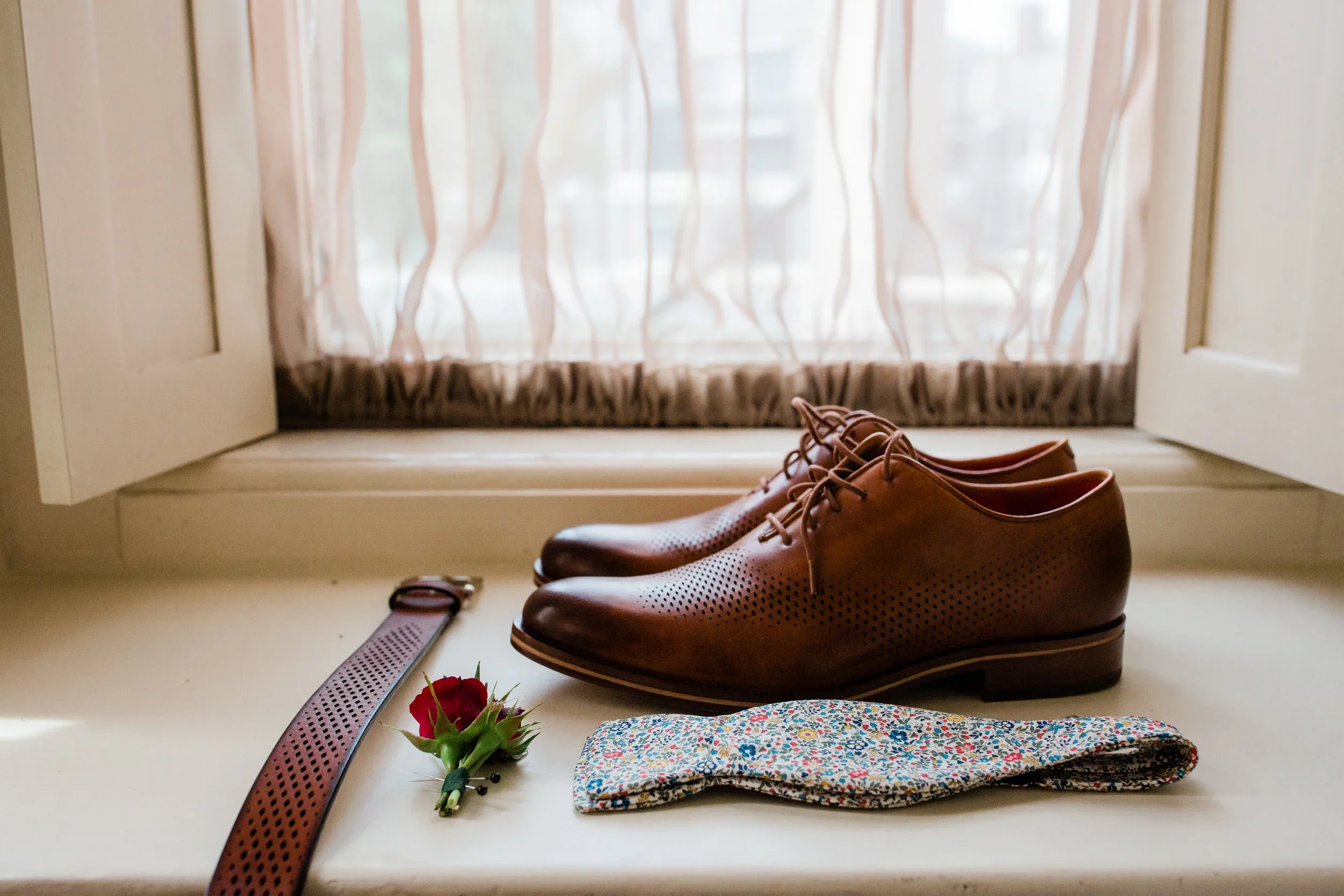 Brown leather dress shoes on a windowsill with a floral-patterned tie, a red rose boutonniere, and a brown leather belt, with a window with light curtains behind.