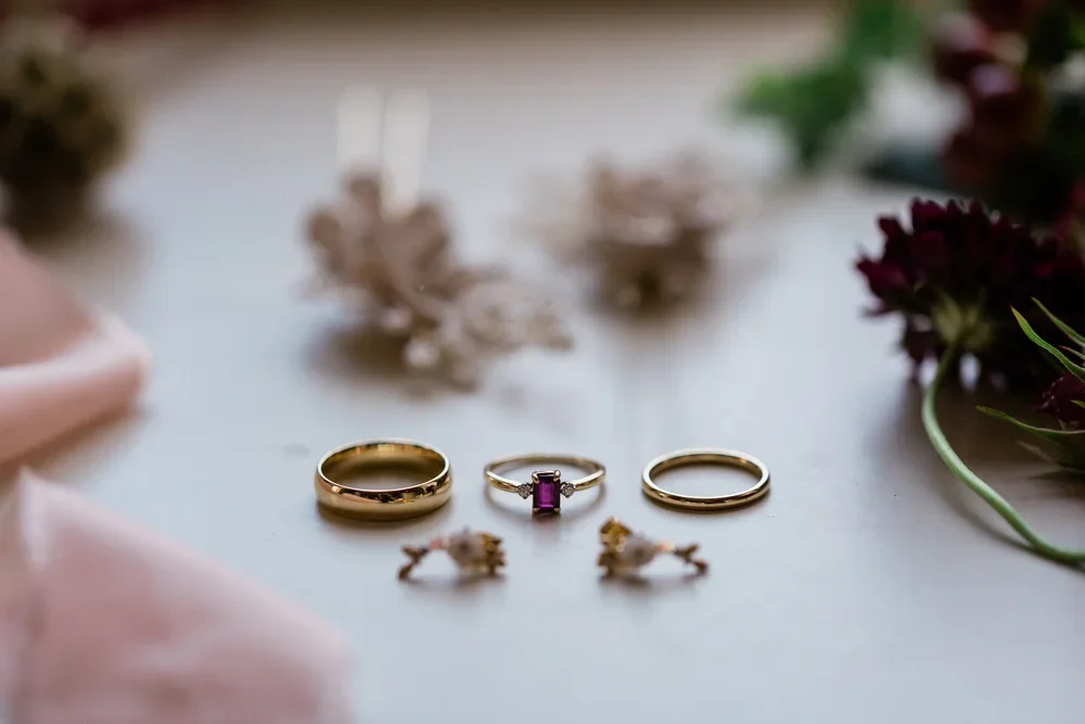 Wedding rings, an engagement ring with a pink gemstone, and earrings on a white surface with flowers and decorations in the background.