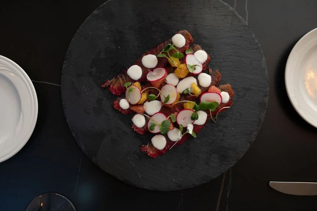 A black slate plate with a dish of seared beef, radish slices, microgreens, and white foam or sauce dots, on a black surface with white side dishes and a knife visible.