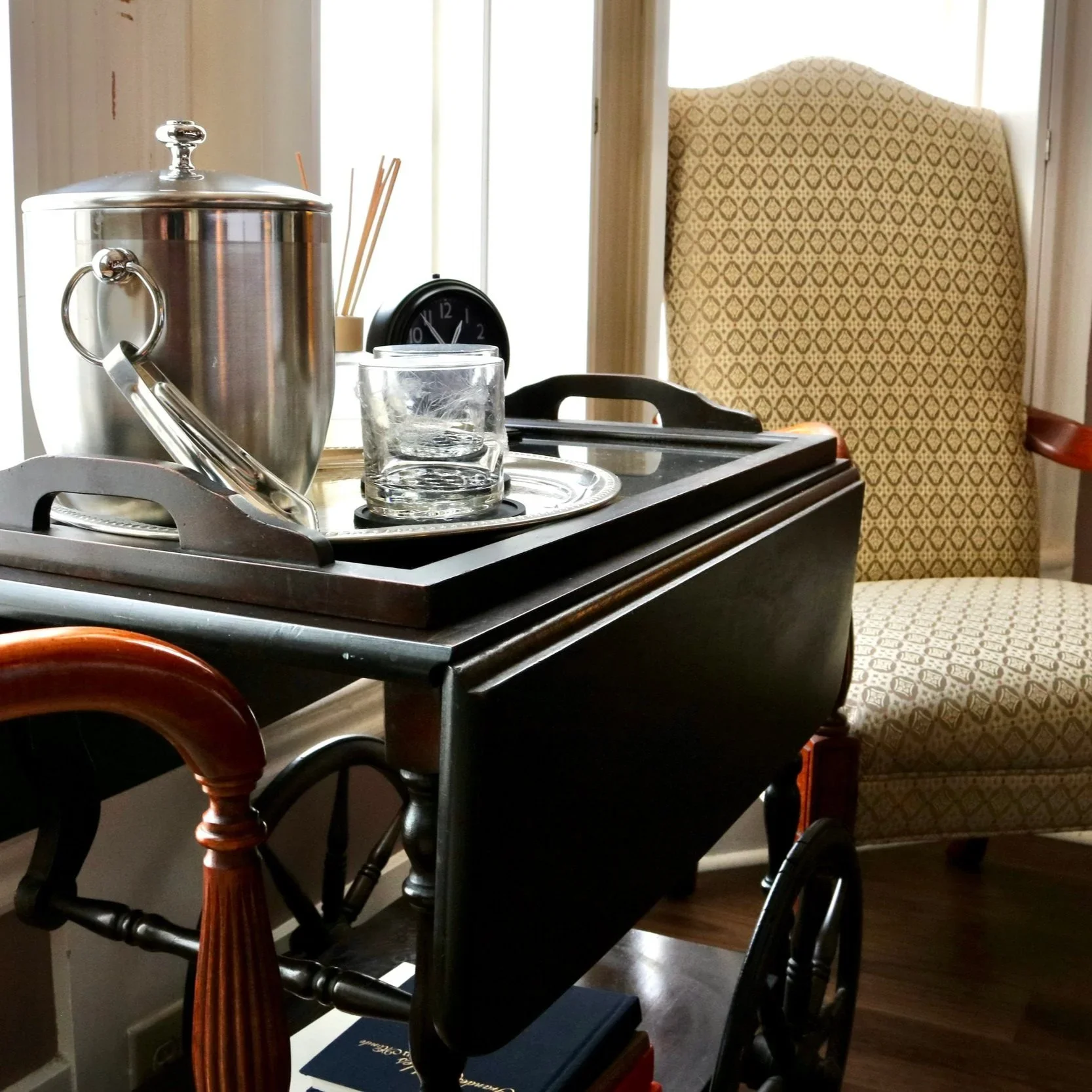 A table with a silver food warmer, a glass tumbler, a clock, and some books, with patterned chairs and a curtain in the background.