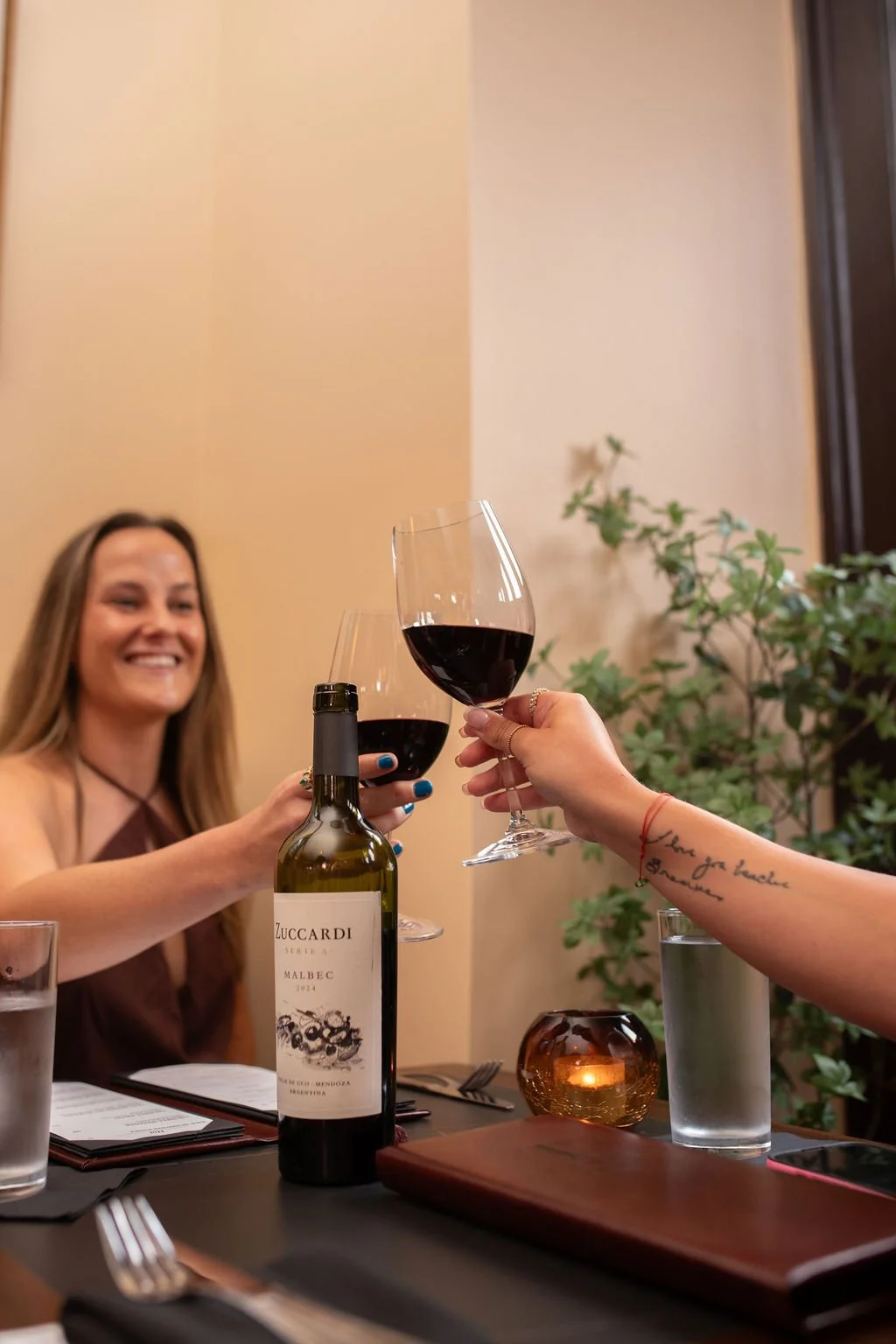 Two women clinking glasses of red wine during a celebration at Table 1888, with a bottle of Zuccardi Malbec on the table, in a warmly lit restaurant setting with plants and candles.