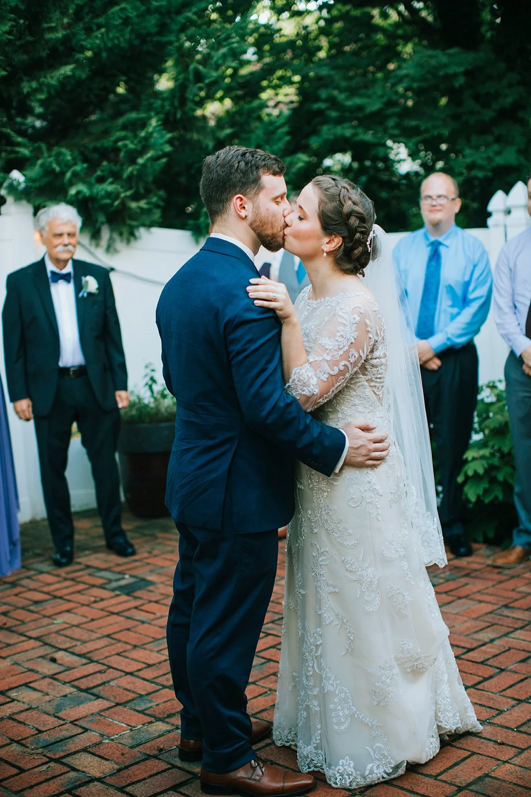 A bride and groom kiss during their wedding ceremony outdoors at Table 1888, with guests in the background and greenery and a white fence behind.